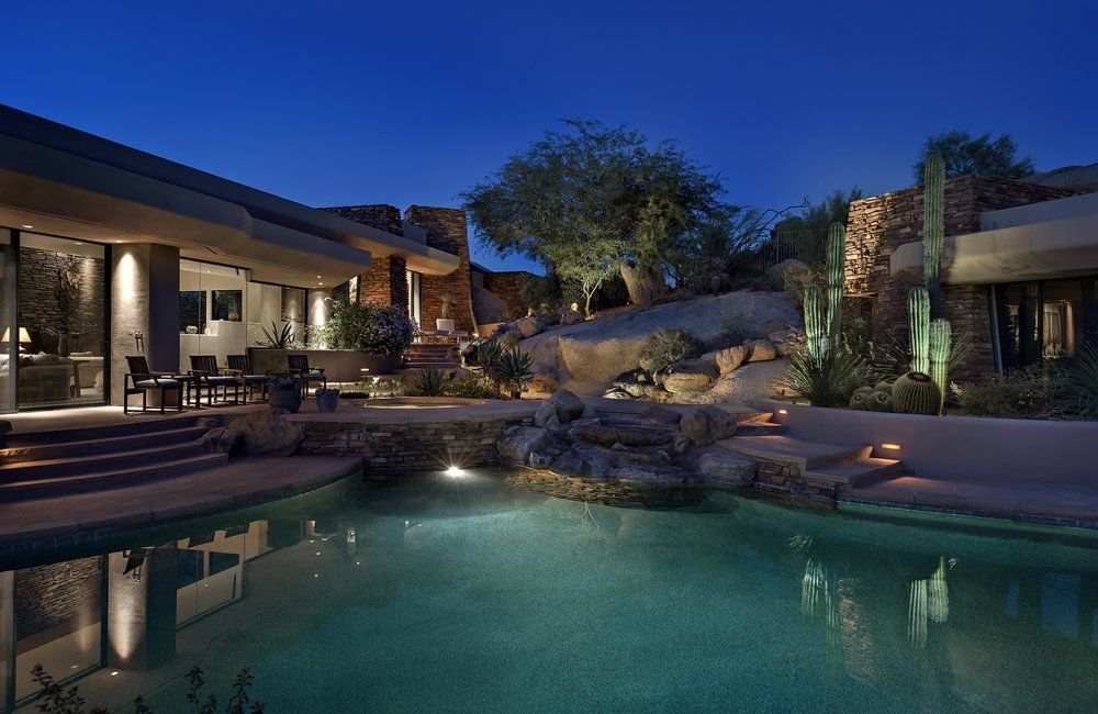 Night view of a luxury home with a pool, waterfalls, and desert landscaping.