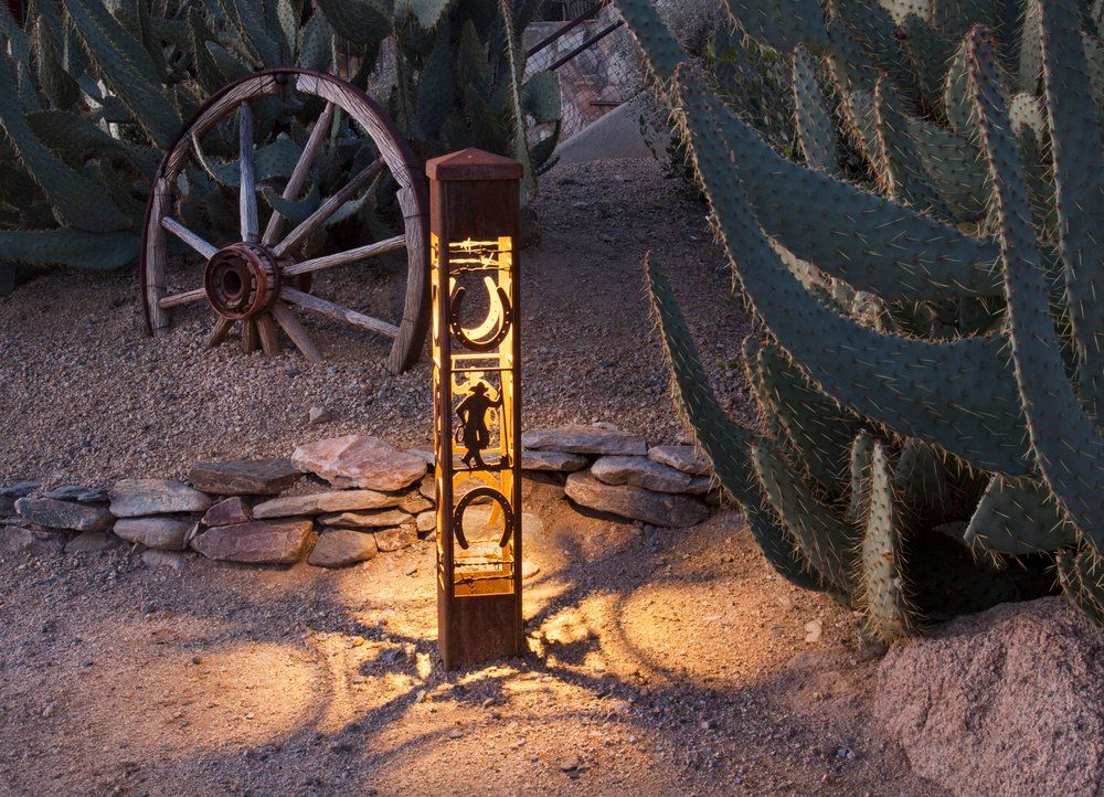 Rustic lighted post with cowboy, moon, and horseshoe cutouts. Beside a cactus and wagon wheel.