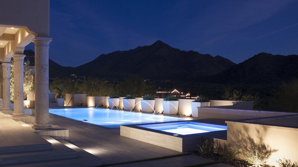 Night view of a lit pool and spa on a patio, with mountains in the background.