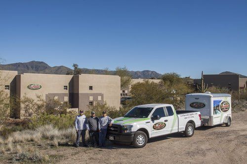 Three men standing by a truck and trailer with company branding in front of a building in a desert landscape.