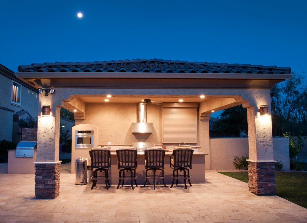 An outdoor kitchen with bar stools under a covered patio at night, illuminated by warm recessed and wall-mounted lights.