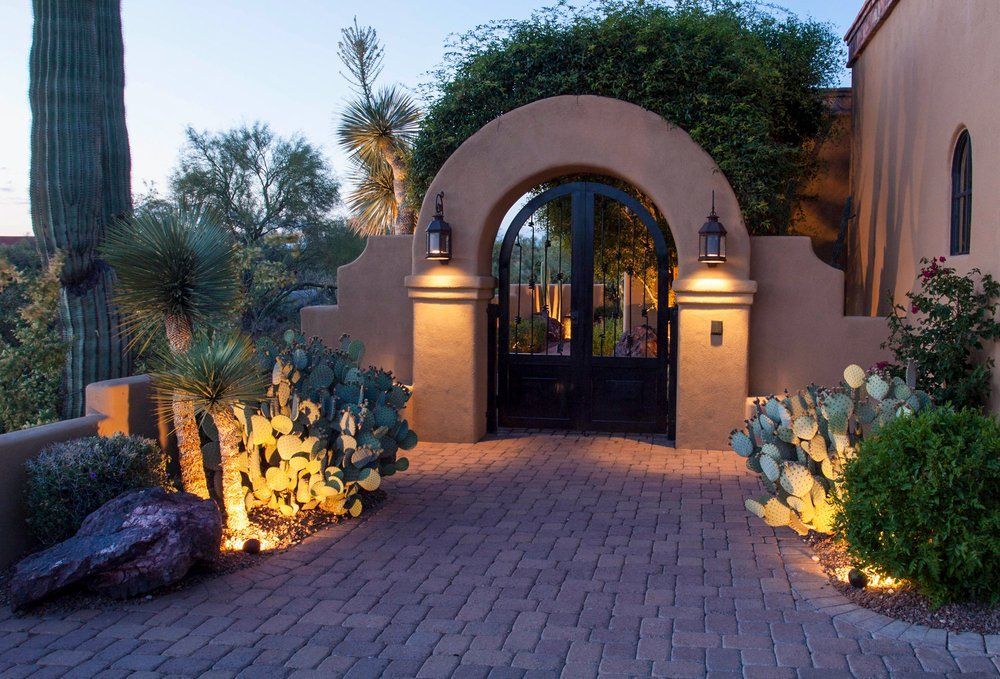 Brick pathway leads to arched black iron gate, illuminated by warm lighting, flanked by cacti and tan stucco walls.