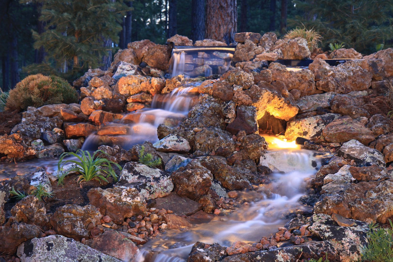 A rock garden waterfall flows into a stream, illuminated by warm lights at dusk among trees.