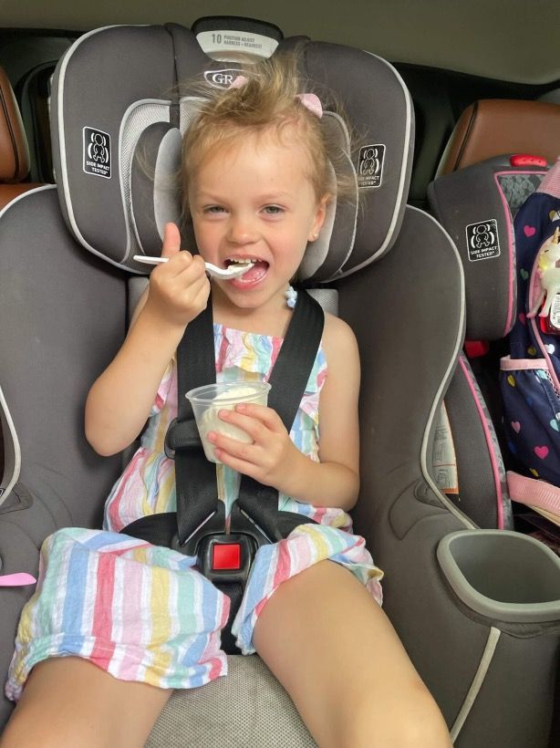 A little girl is sitting in a car seat eating ice cream.