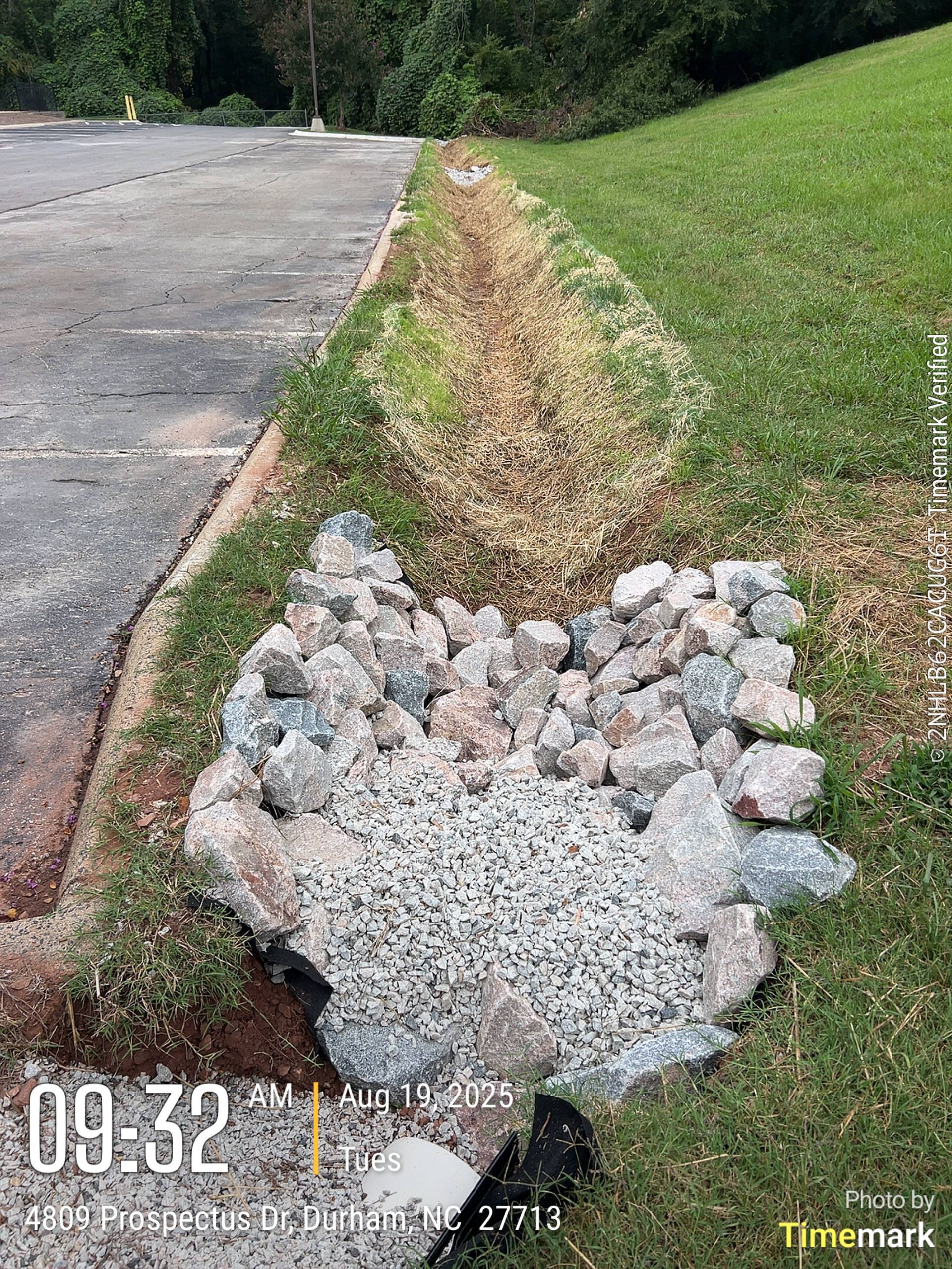 Rocks in a gravel-lined drainage ditch next to a grassy hill and parking lot.