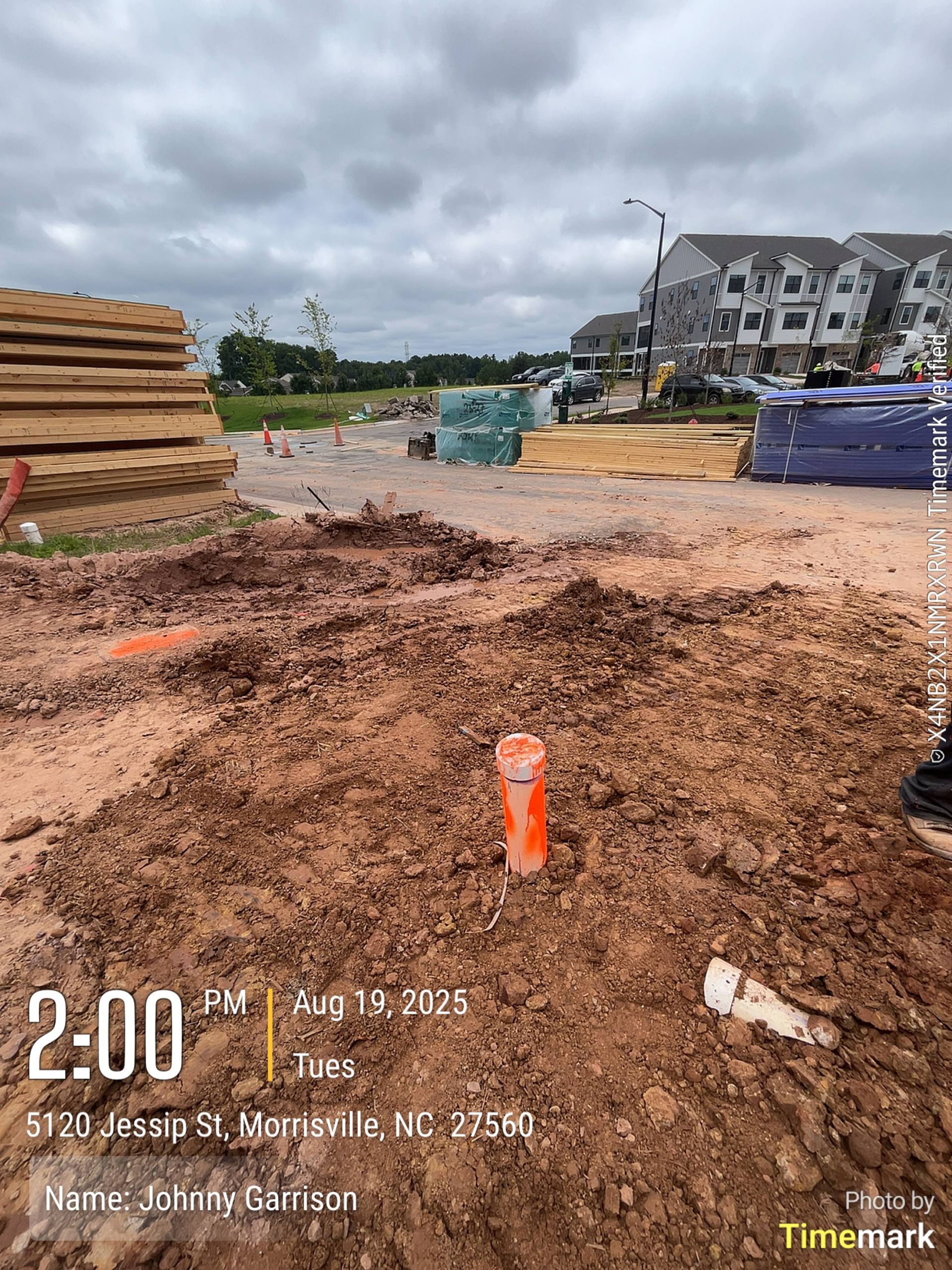 Construction site with dirt, orange marker, lumber, and buildings under cloudy sky.