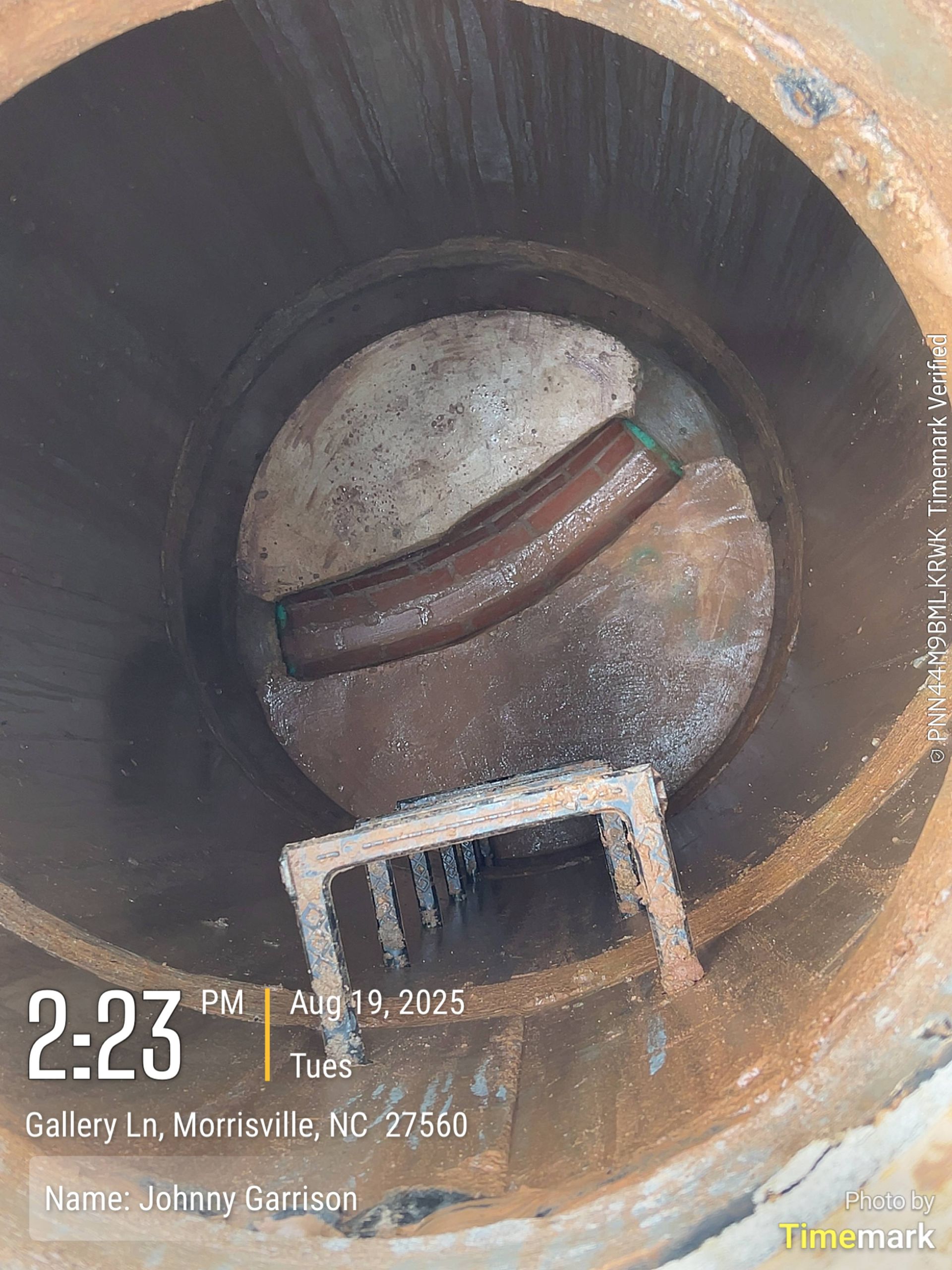 View inside a dark, rusty manhole with a curved pipe and a small metal grate.