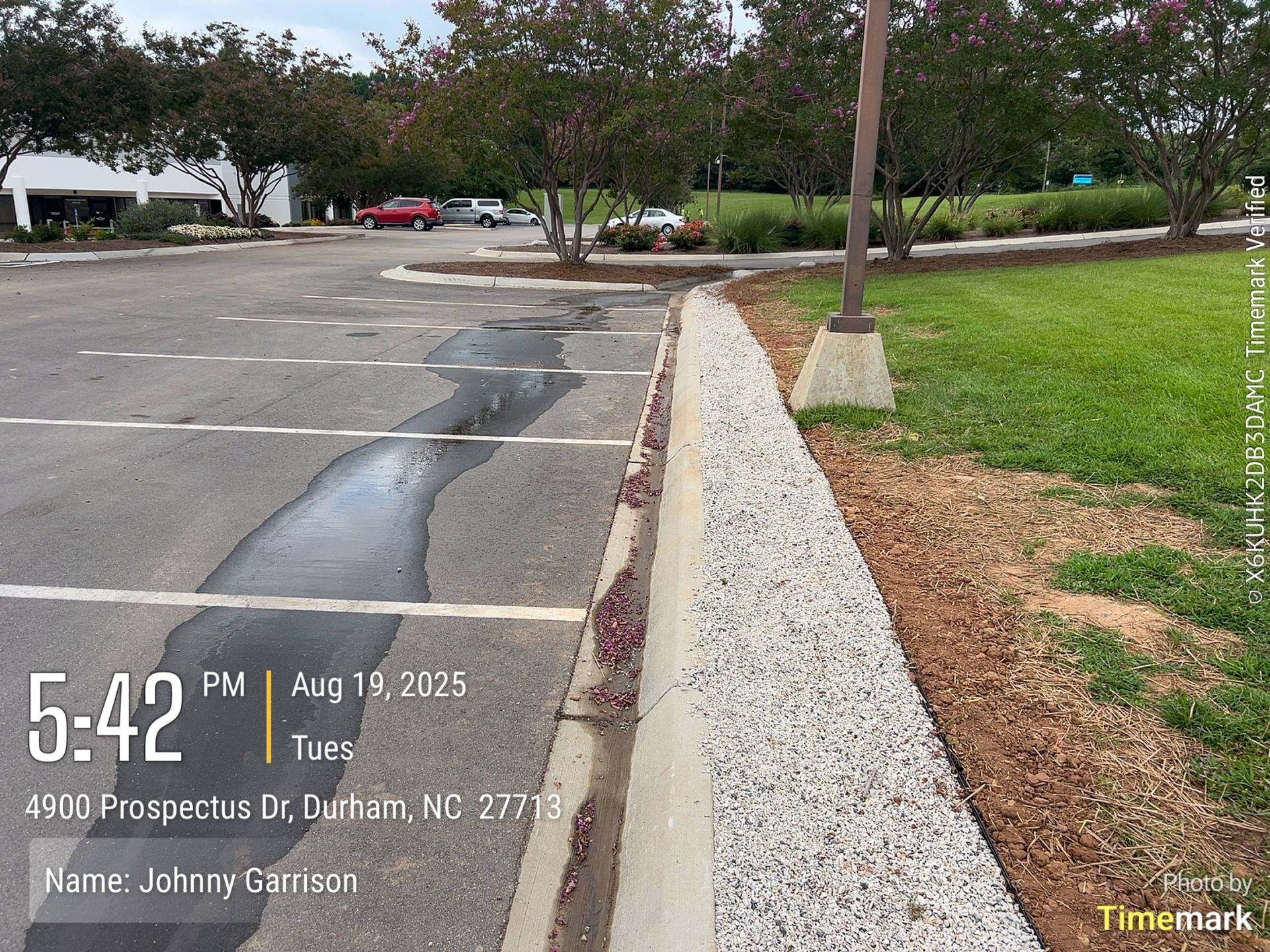Parking lot with gravel-lined curb, sidewalk, and grass; building and cars in background.