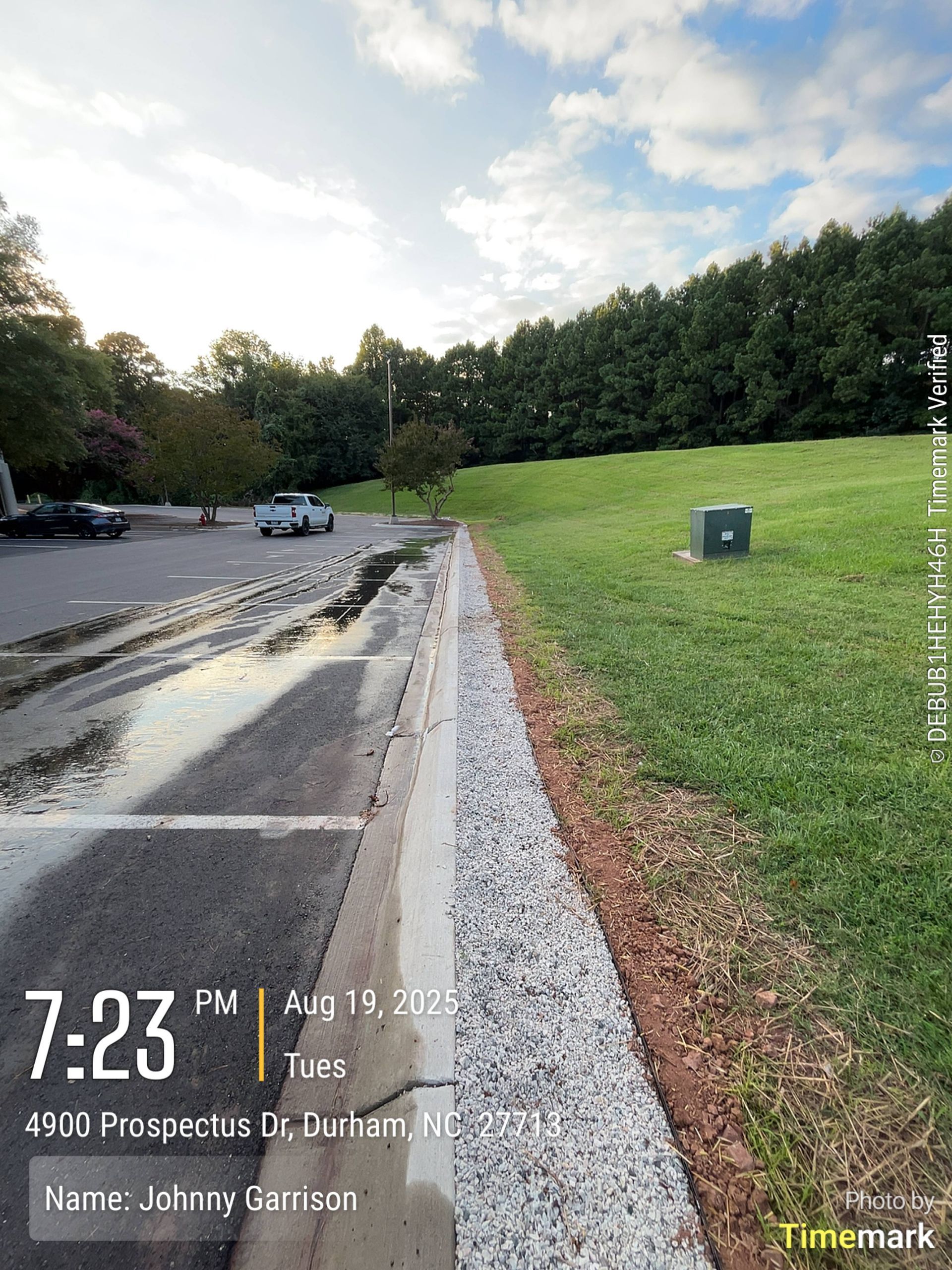 A parking lot next to a grassy area with a curb and a drainage system; the sky is partly cloudy.