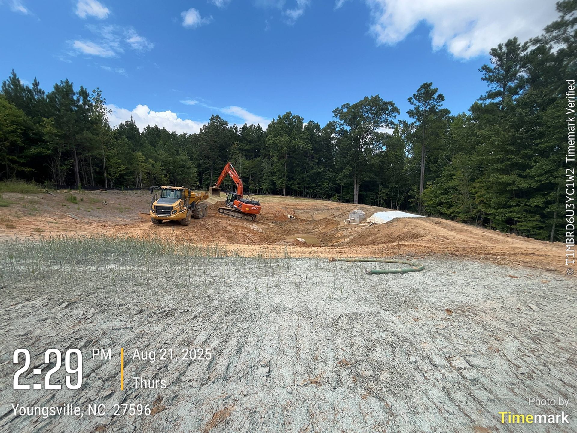 Construction site with an excavator and a small bulldozer on a dirt lot, under a blue sky with trees in the background.