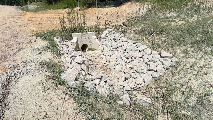 Dirt road with a concrete drainage structure, surrounded by gravel and grass. Trees in background.