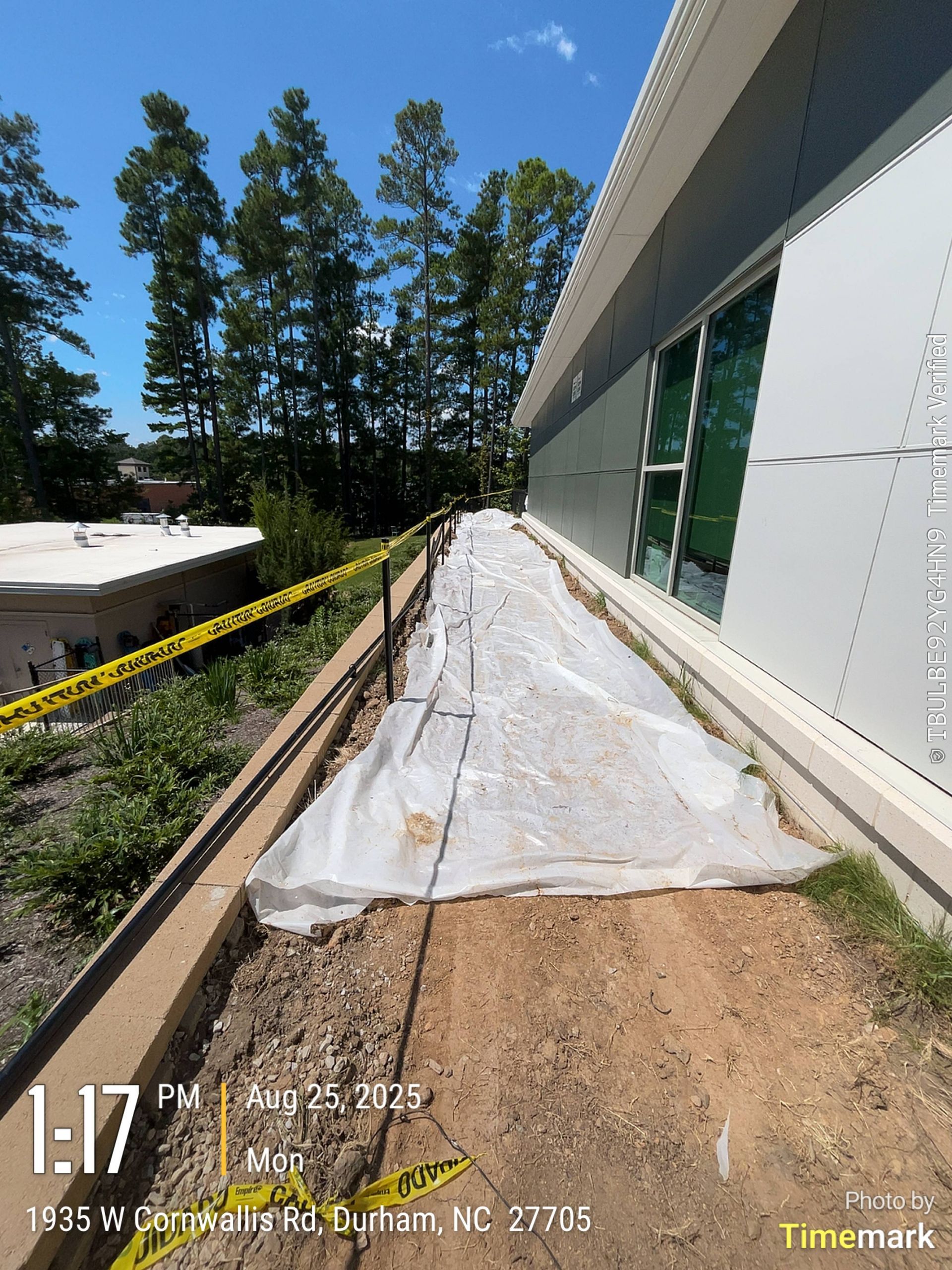 Construction site with plastic covering walkway next to modern building. Yellow caution tape, trees.