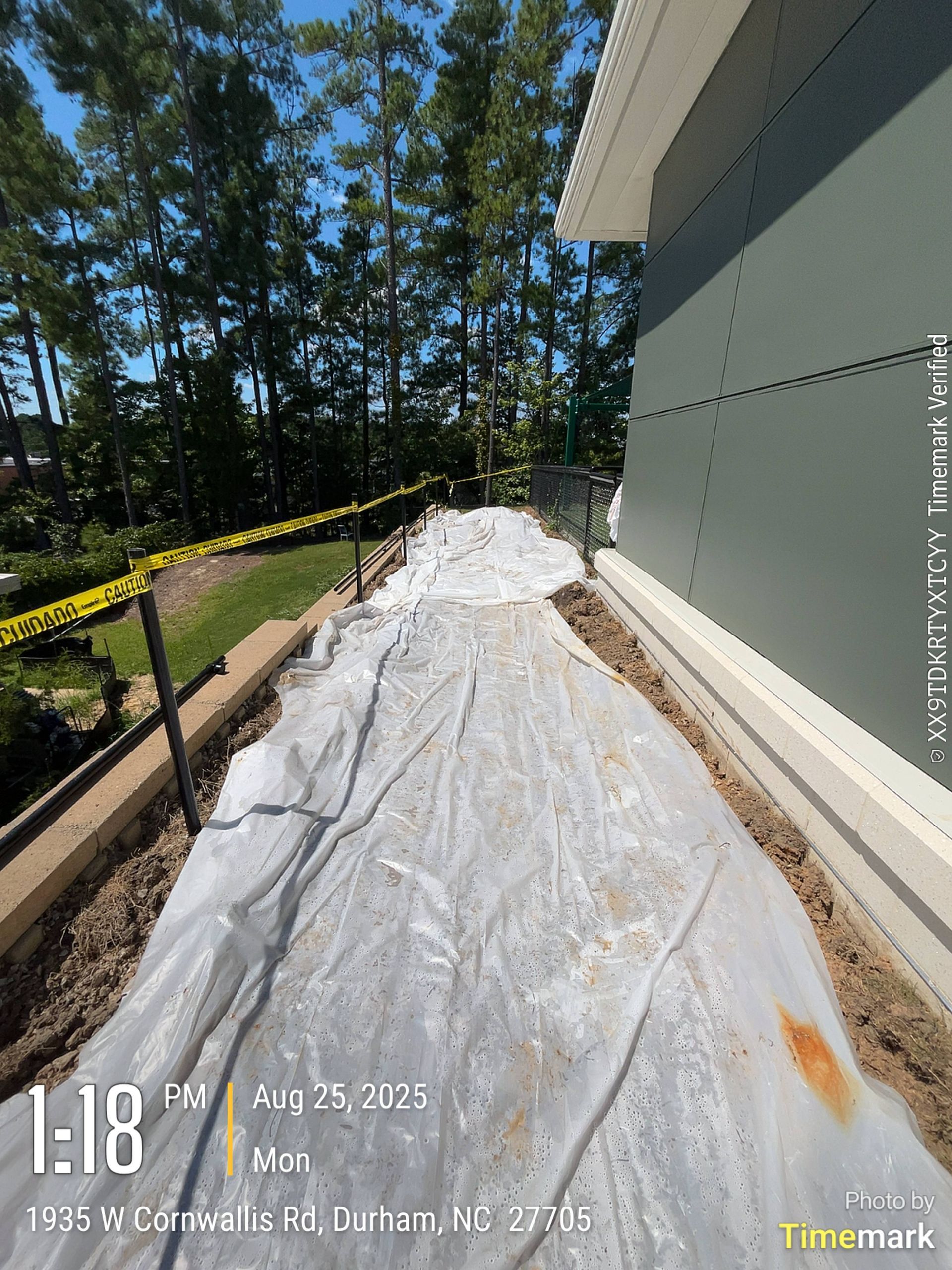 A plastic sheet covers a narrow path next to a building, bordered by soil and caution tape; Durham, NC.