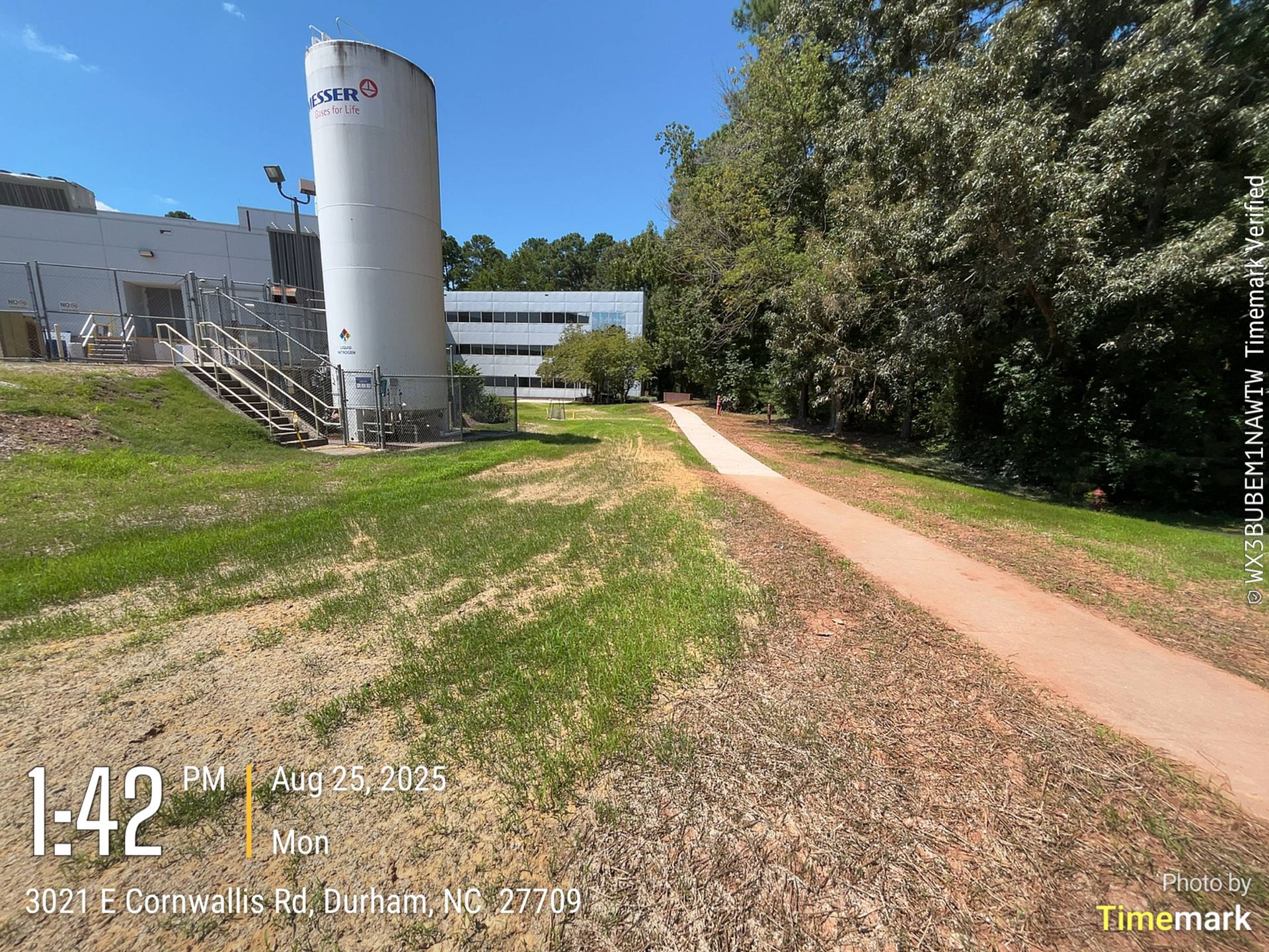 A paved path winds past a white industrial building and tall silo, adjacent to a grassy area with trees.