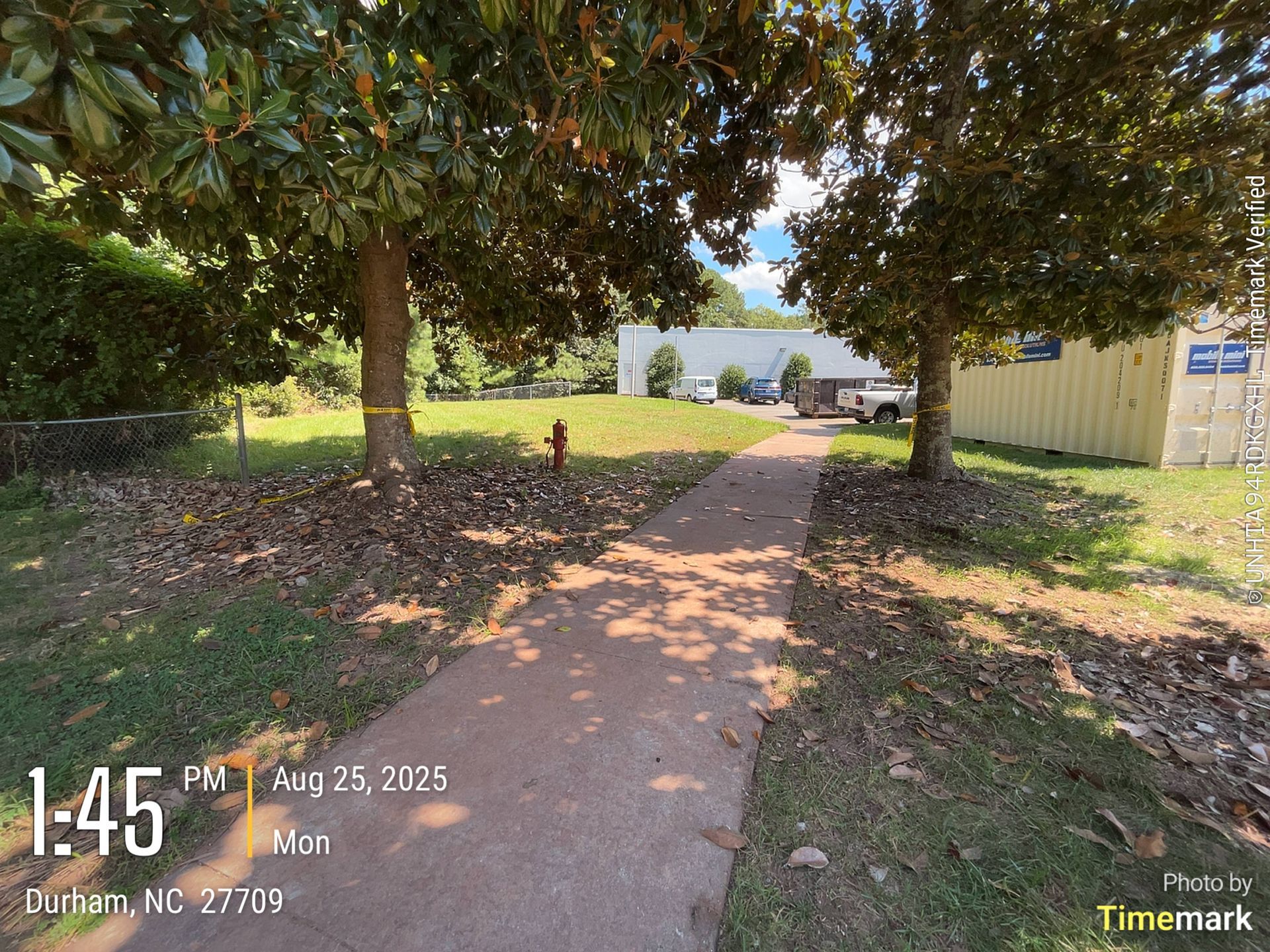 A paved path leads past trees toward buildings in Durham, NC, on a sunny afternoon.