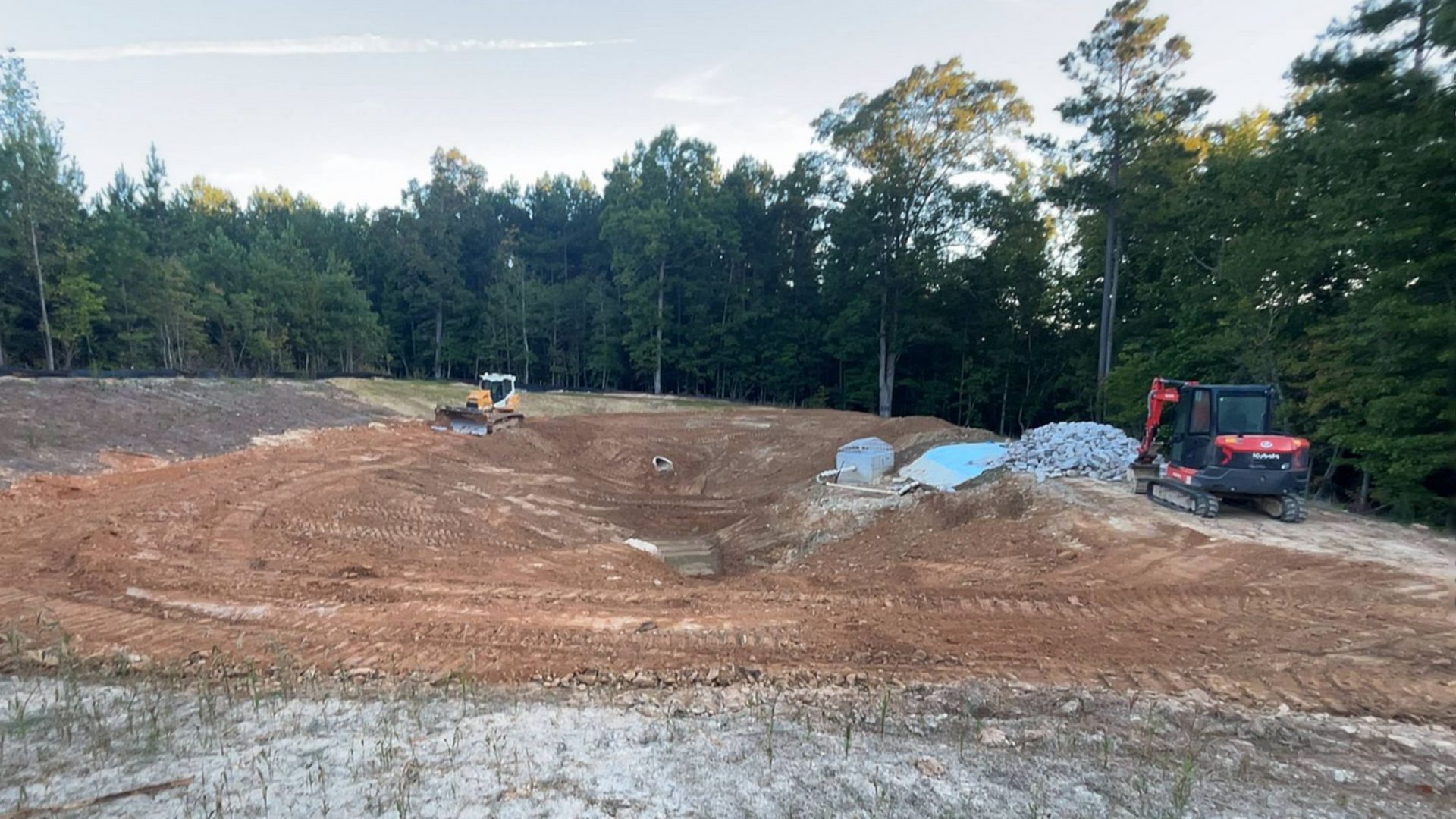 Construction site with two excavators, dirt mounds, and trees in the background.