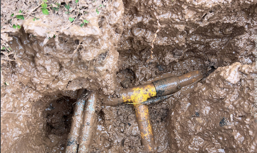 Yellow-handled tool partially buried in muddy soil, alongside exposed metal pipes.