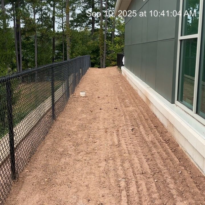 A dirt path between a black fence and a building, with tire tracks. Trees are in the background.