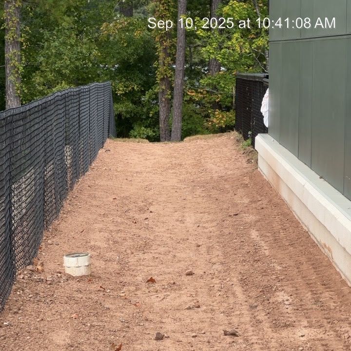 A dirt path alongside a building and fence, with trees in the background.