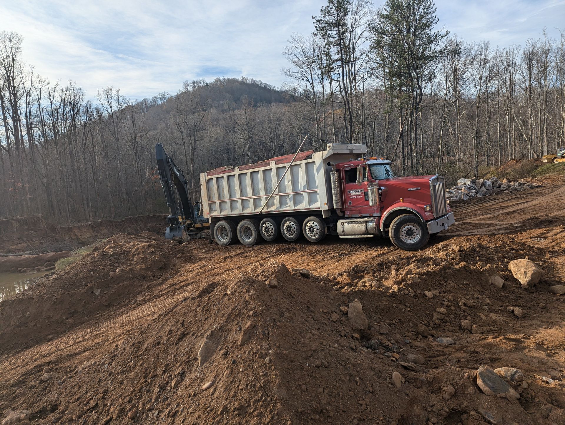 Red dump truck hauling dirt on a construction site. Hills and trees are in the background.