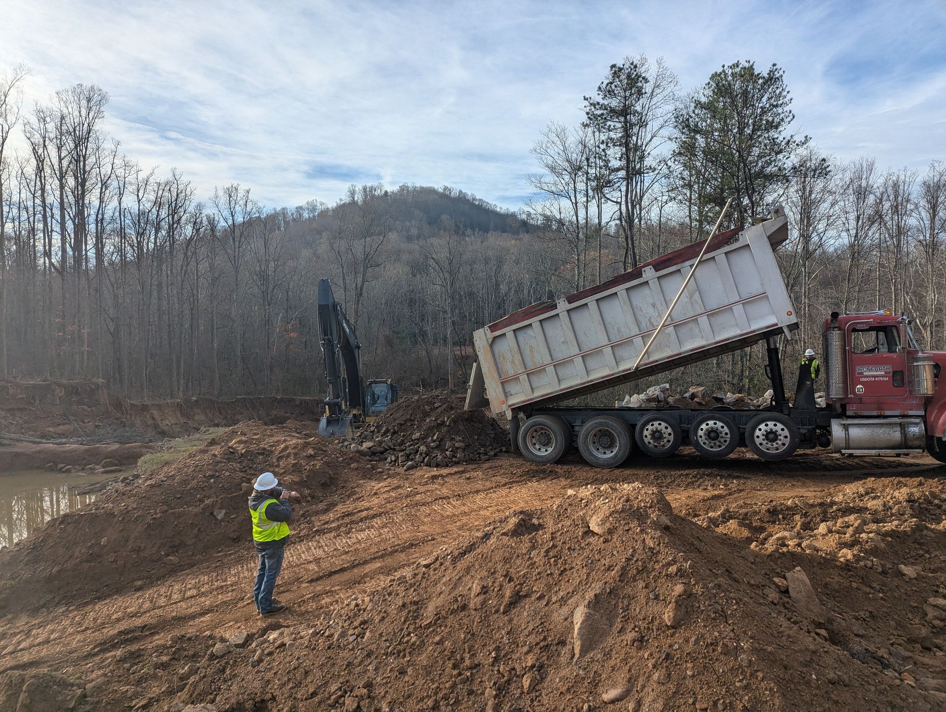 Construction site: Dump truck unloading dirt, excavator, worker in safety vest, trees, mountain in background.