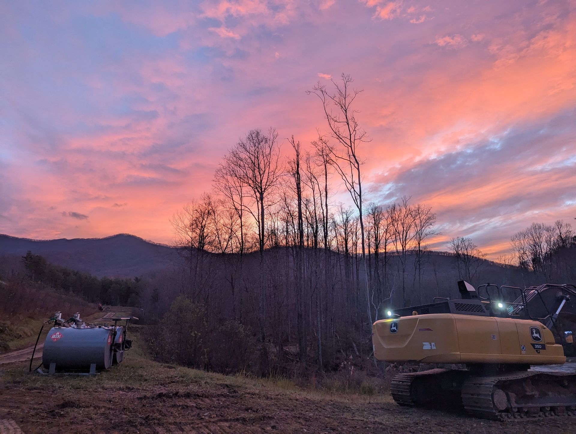 A construction site at sunrise. A yellow excavator and a fuel tank are on a dirt lot, with a colorful sky.