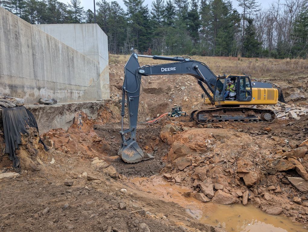 A John Deere excavator digs into dirt near a concrete structure at a construction site in a wooded area.