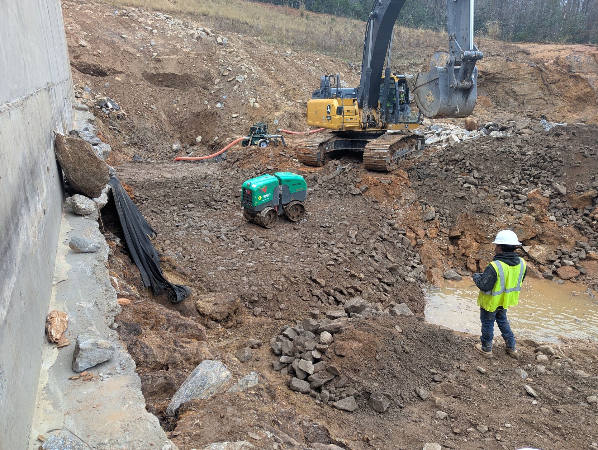 Construction site: excavator, worker in safety vest, generator, and muddy ground.