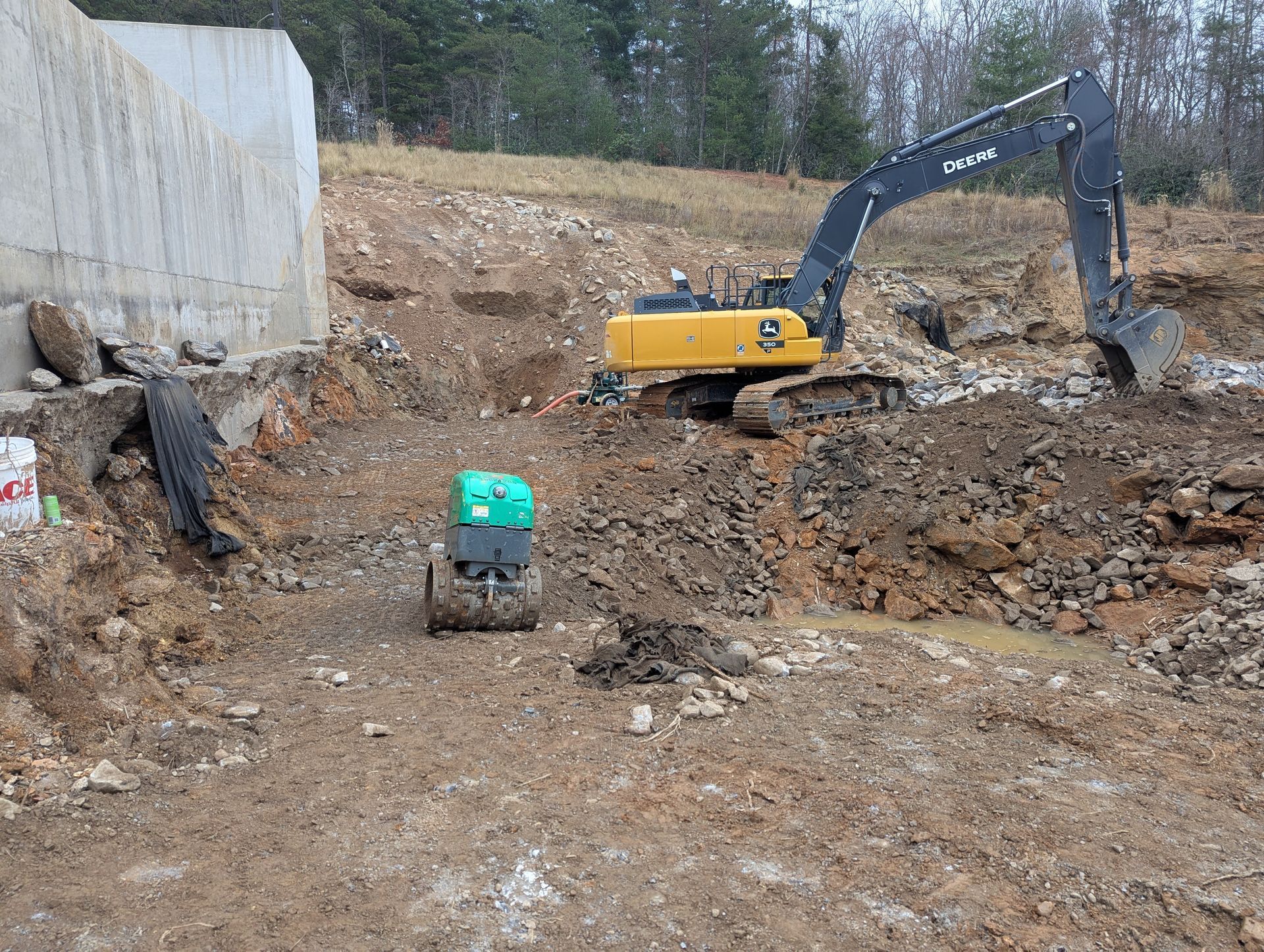 Excavator and small compactor working on a construction site with a concrete structure and earth.