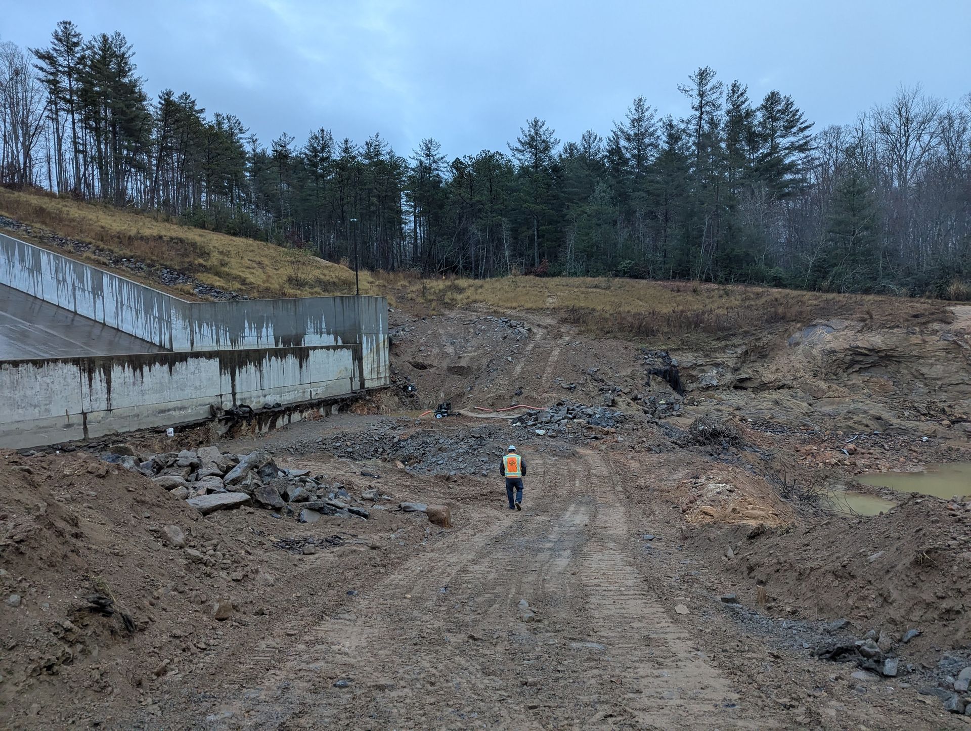 Person in an orange vest walks through muddy construction site near a concrete wall and trees.