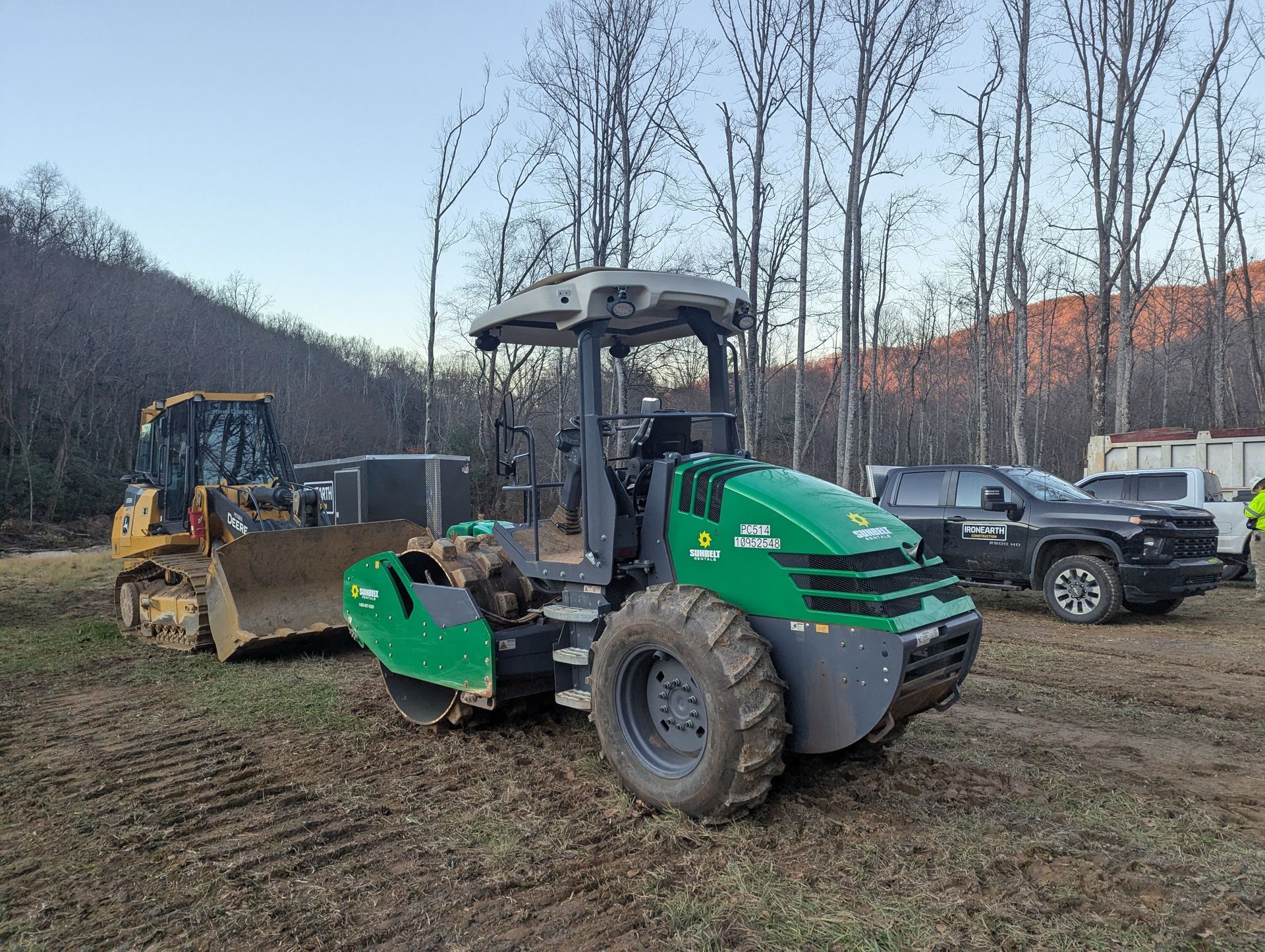 Green and black roller compactor on muddy ground, with bulldozer and truck in the background.