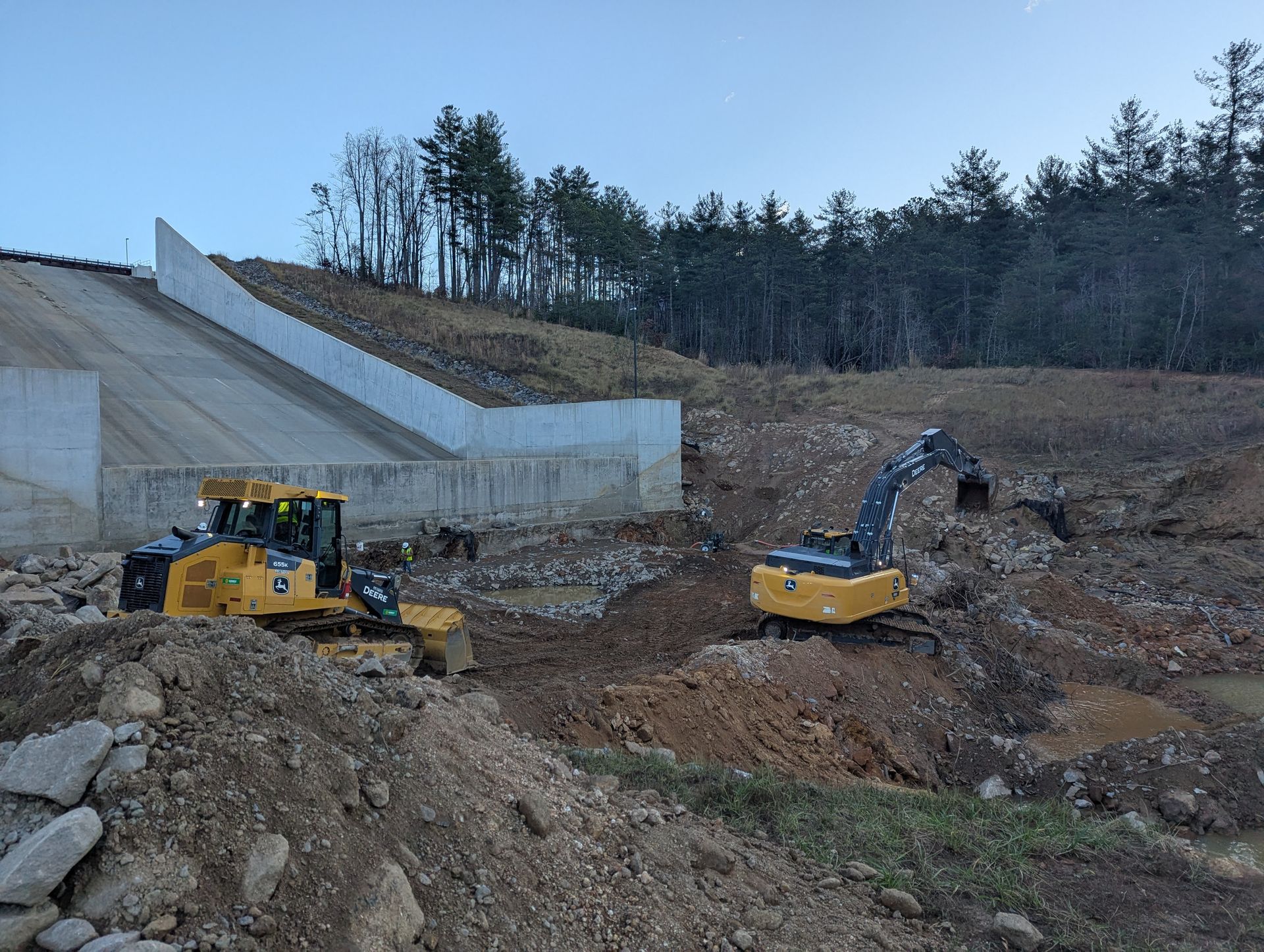 Yellow bulldozer and excavator on a construction site next to a concrete wall and trees.
