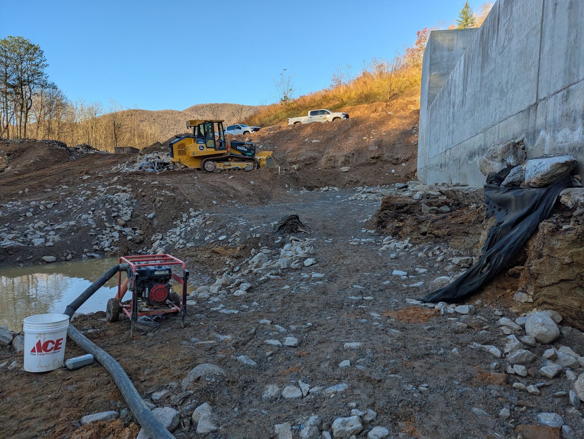 Construction site with bulldozer, concrete wall, and pump near a water source.