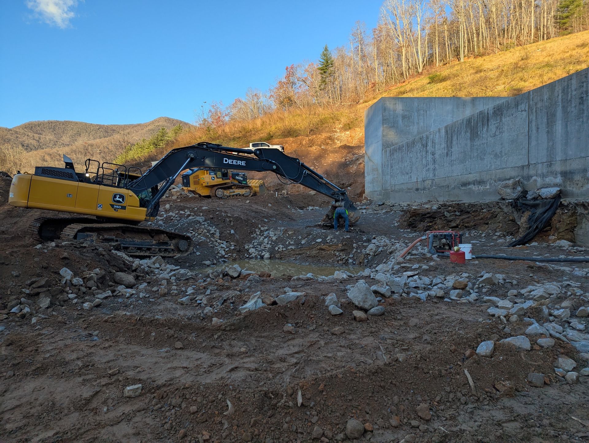 A yellow excavator demolishes concrete near a large wall, mountains in the background.