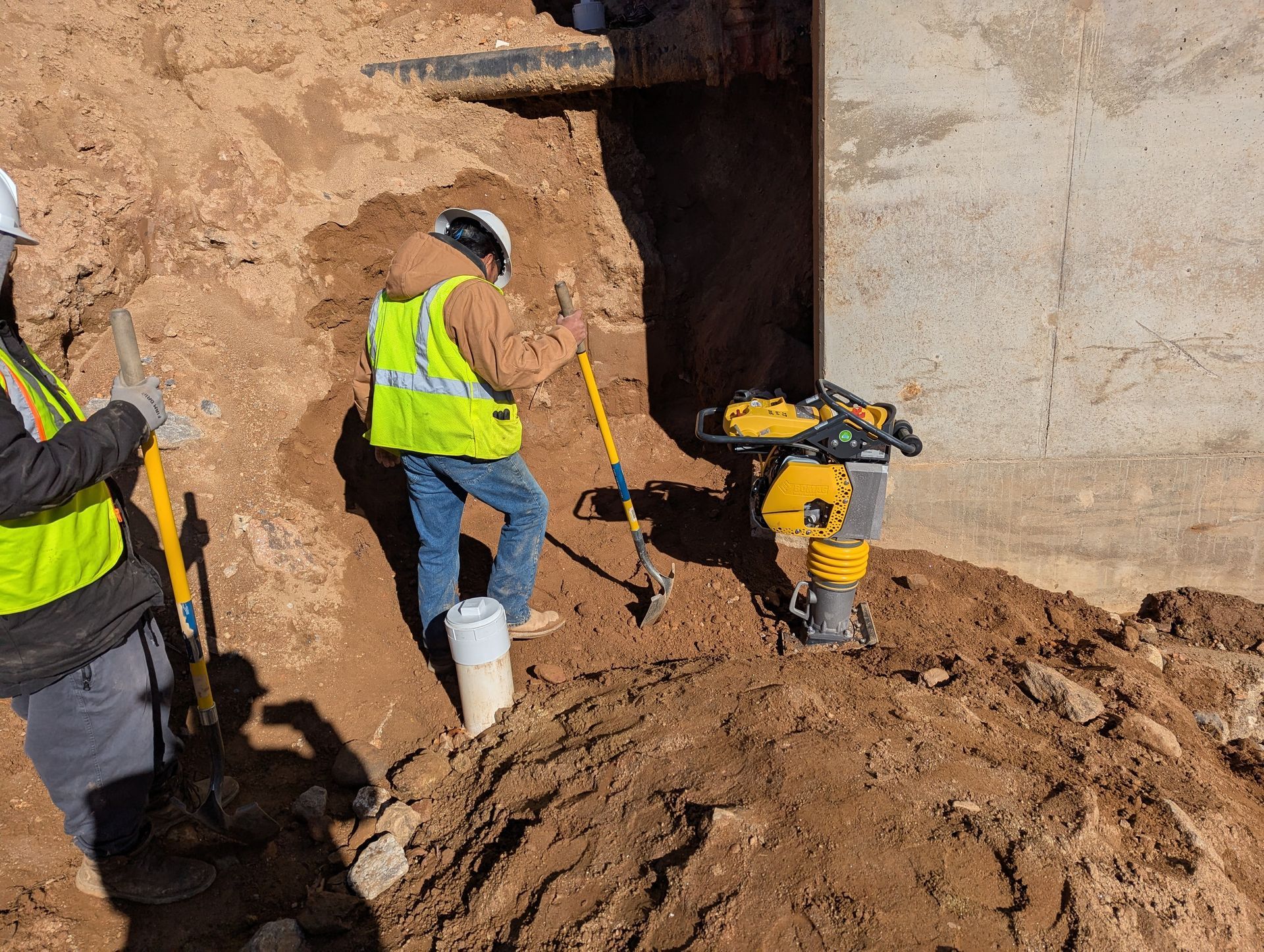 Construction workers excavating earth near a concrete structure, one operating a compactor, others using shovels.
