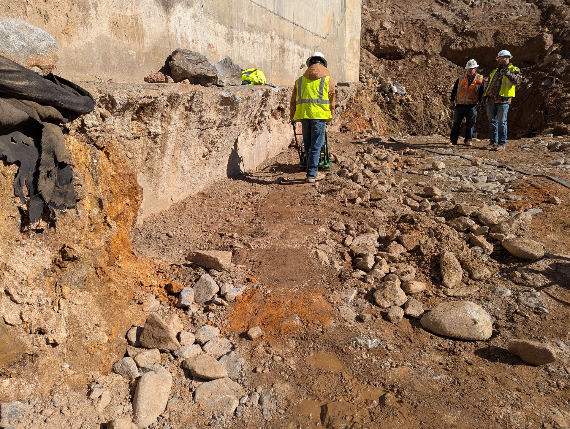Construction workers near a concrete structure on a rocky terrain. One worker in focus, wearing a reflective vest.