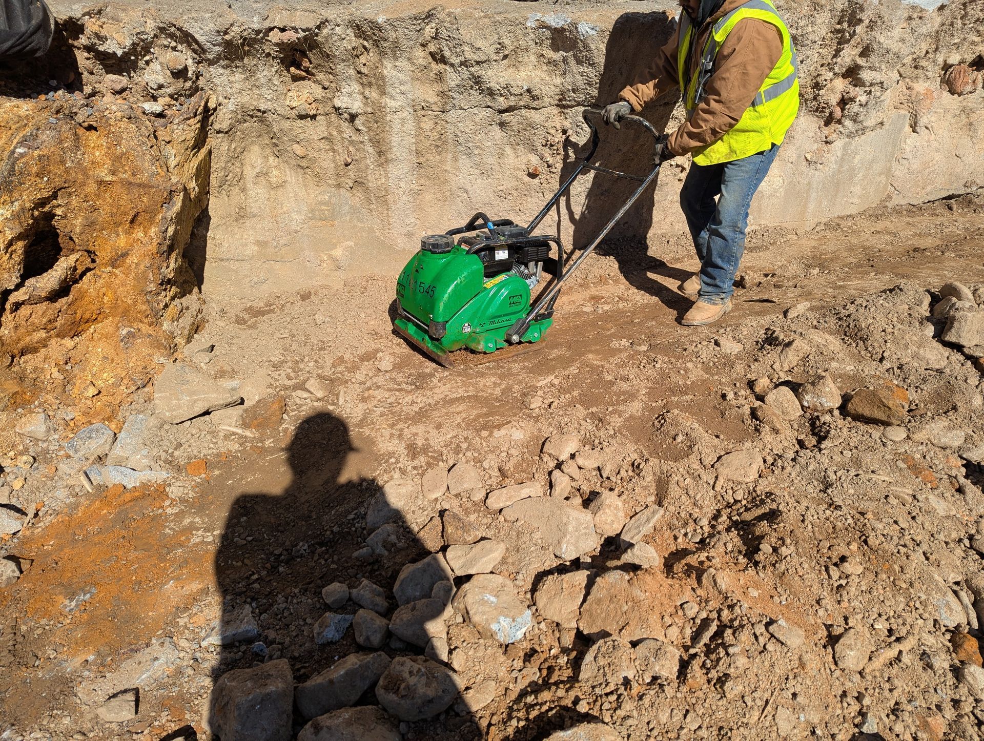 Worker compacting soil in a construction site, wearing safety vest and hard hat.