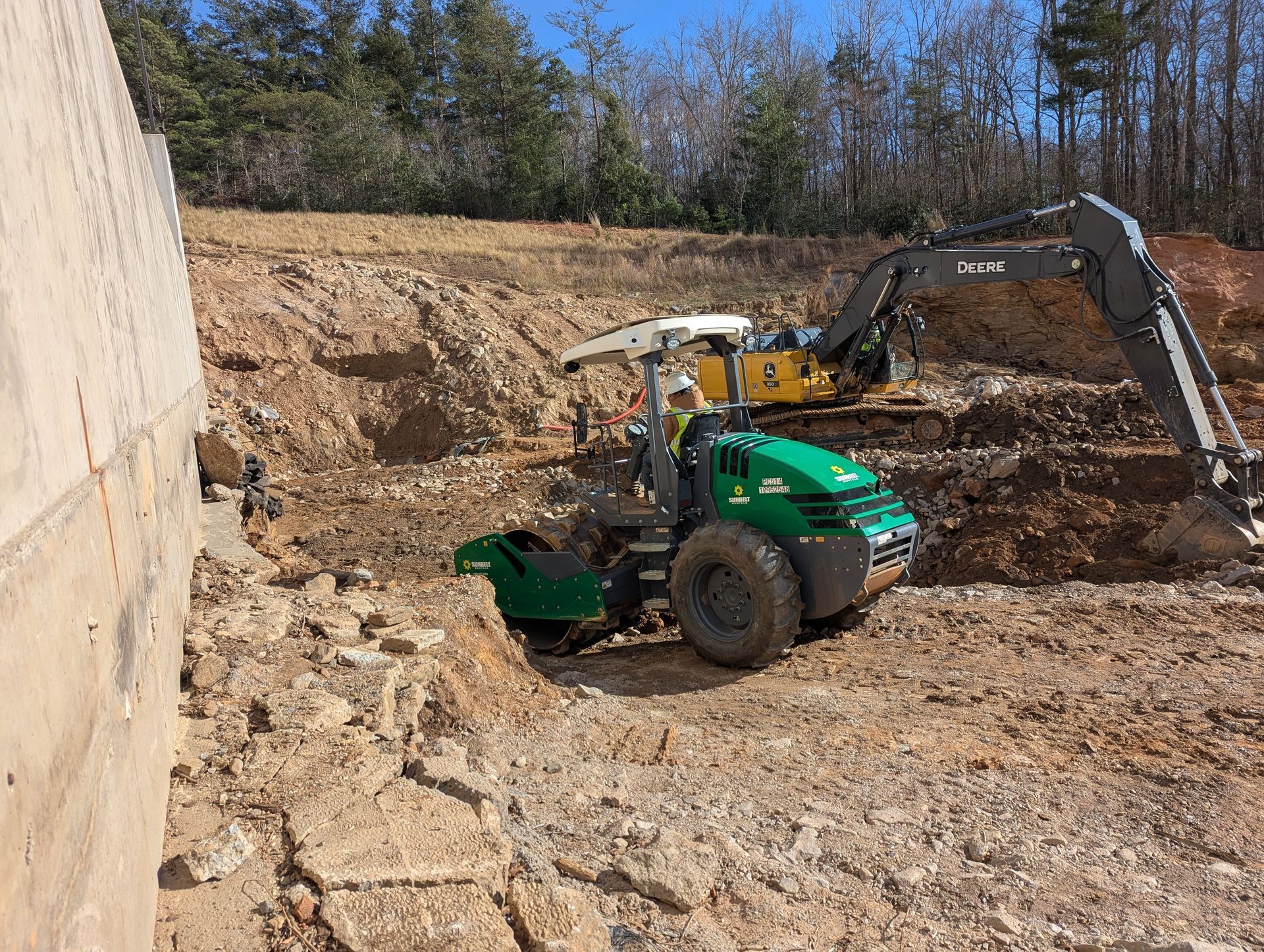 Construction site: Green compactor and yellow excavator working on a hillside near a concrete wall.