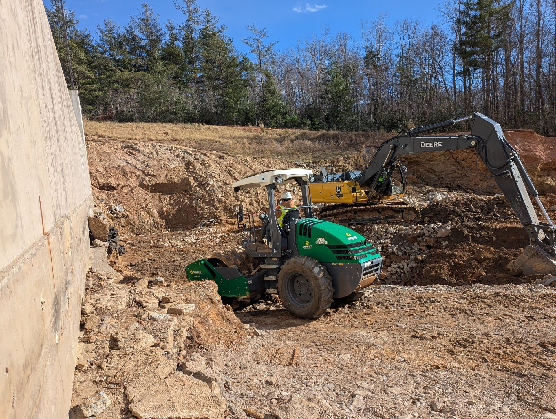 Construction site with excavator and compactor working on a dirt hill, near a concrete structure and trees.