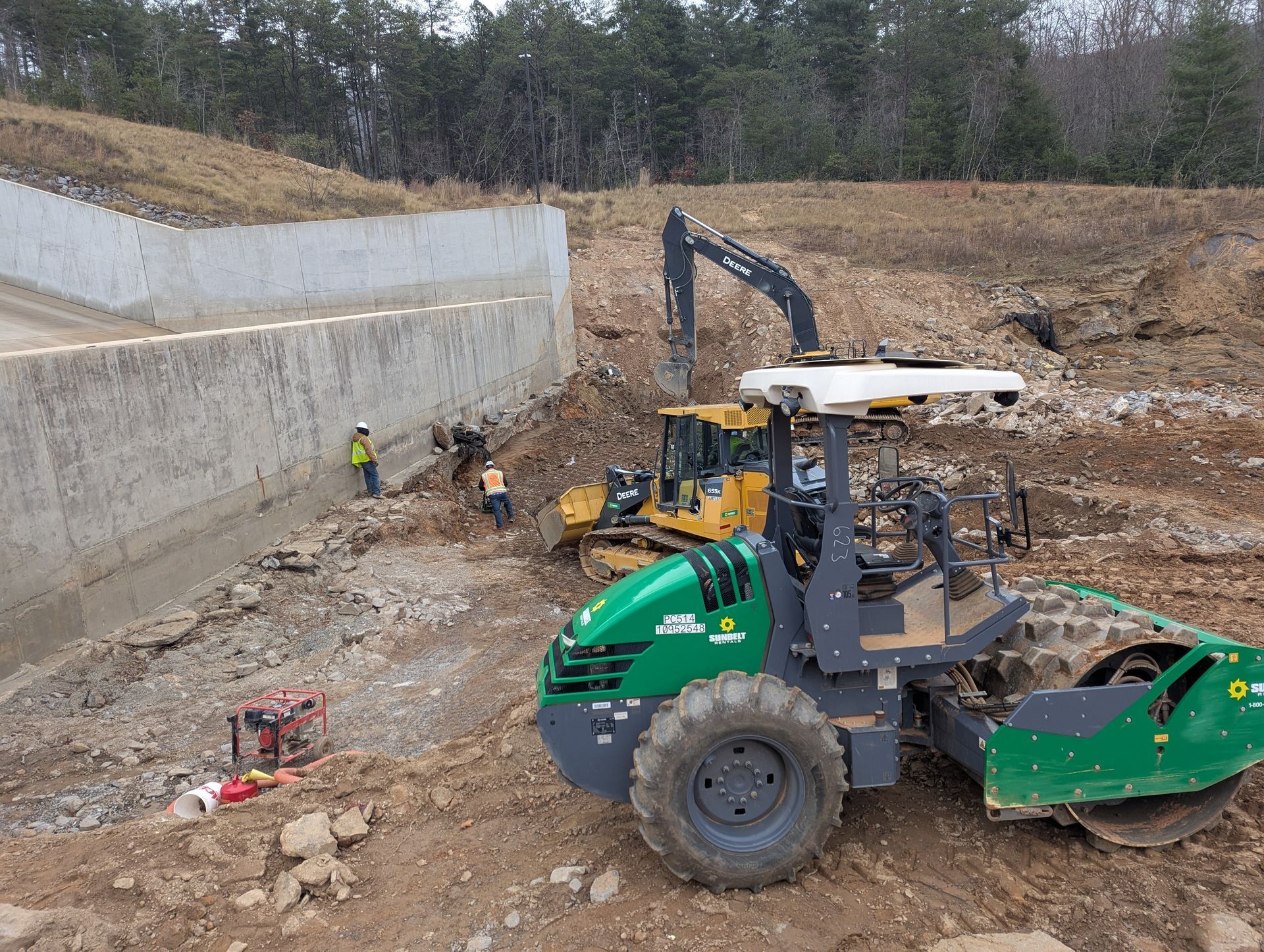 Construction site: green roller, yellow bulldozer, excavator and workers near a concrete wall.