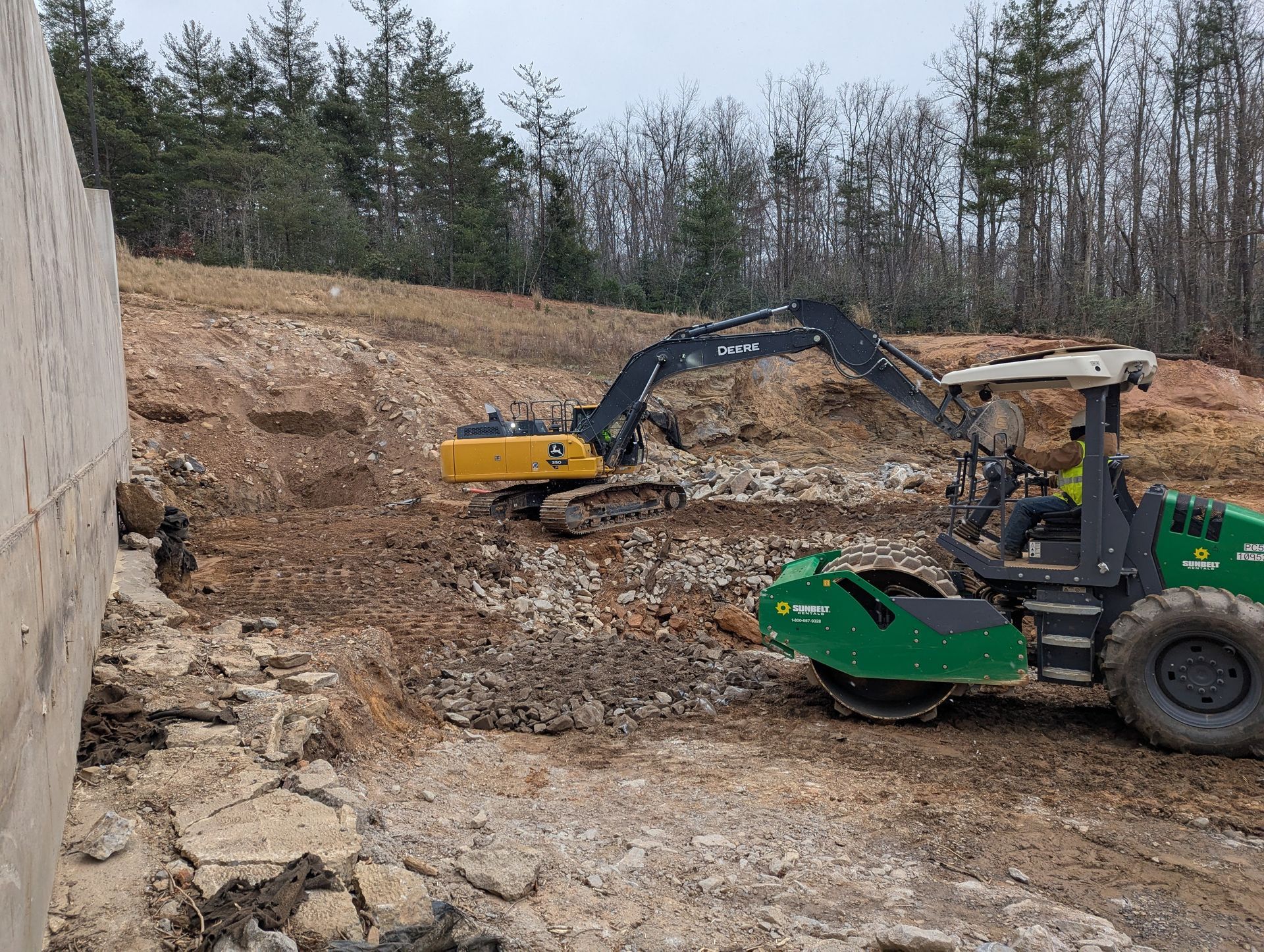 An excavator and a compactor working at a construction site with a concrete wall.