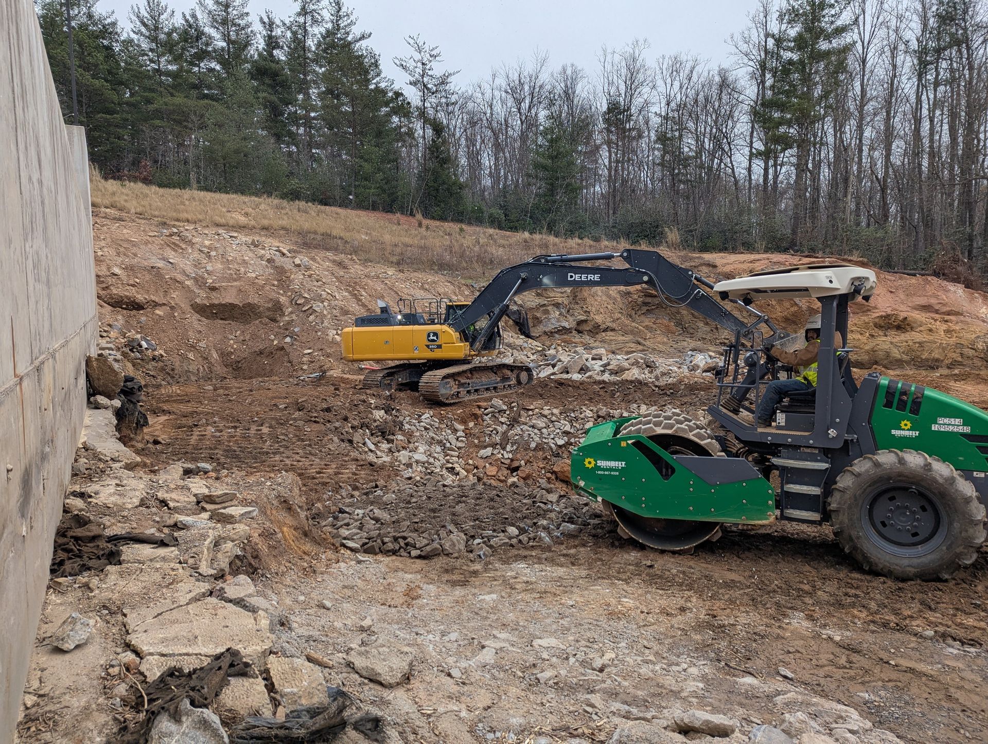 Construction site: Excavator and roller compacting earth near a concrete wall and treeline.