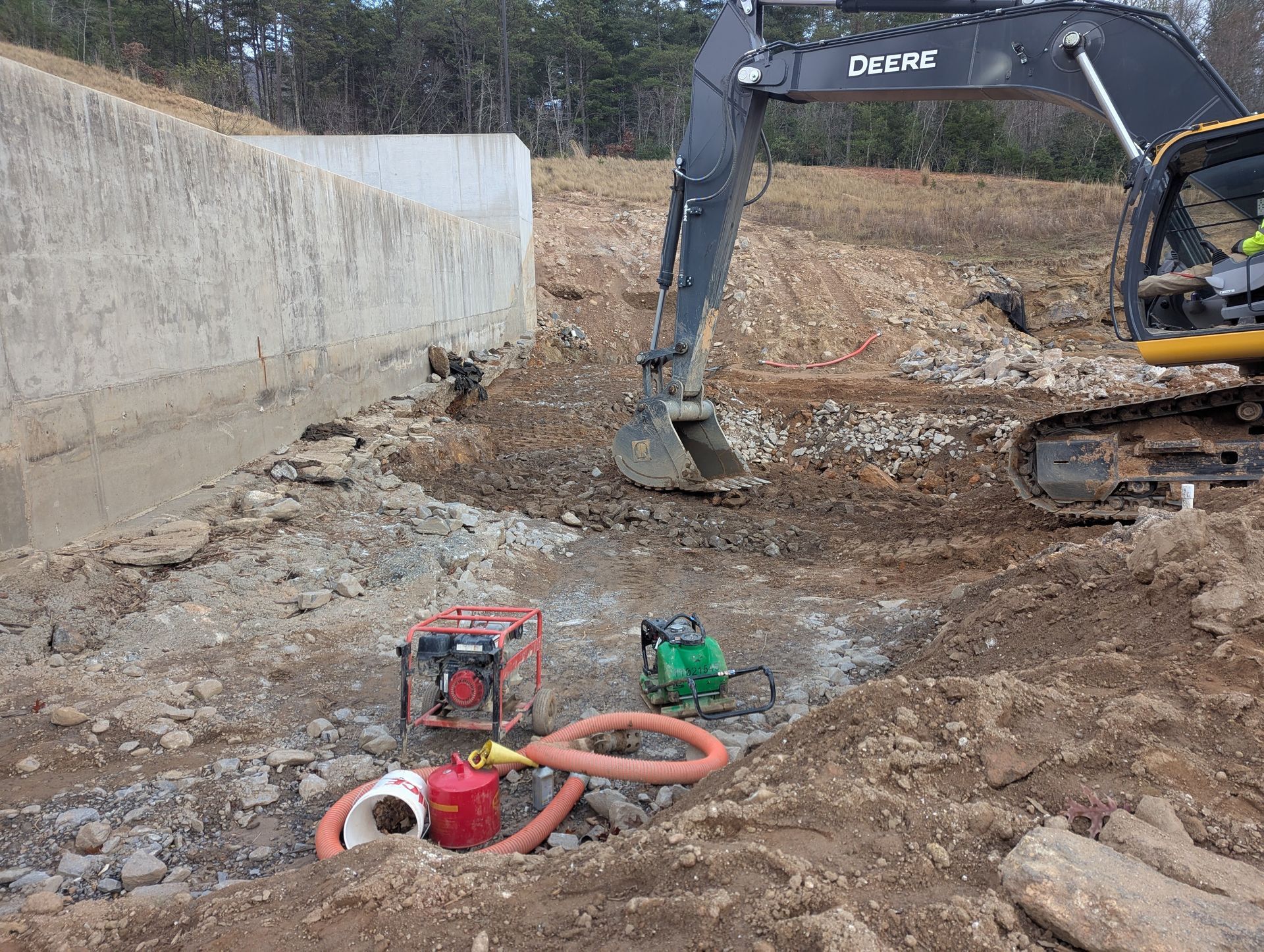 An excavator working at a construction site near a concrete wall; two water pumps and hoses are nearby.