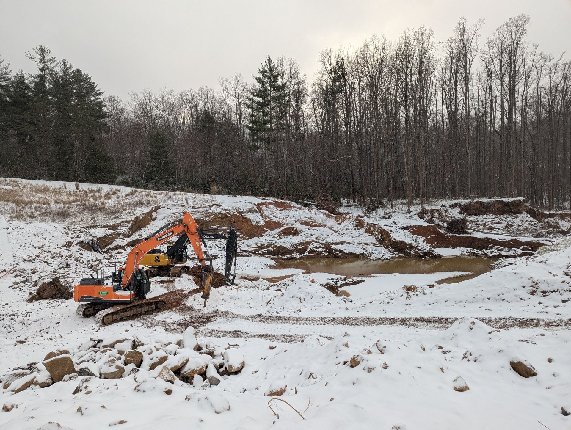 An orange excavator and a person are working on a muddy site covered in snow, with trees in the background.