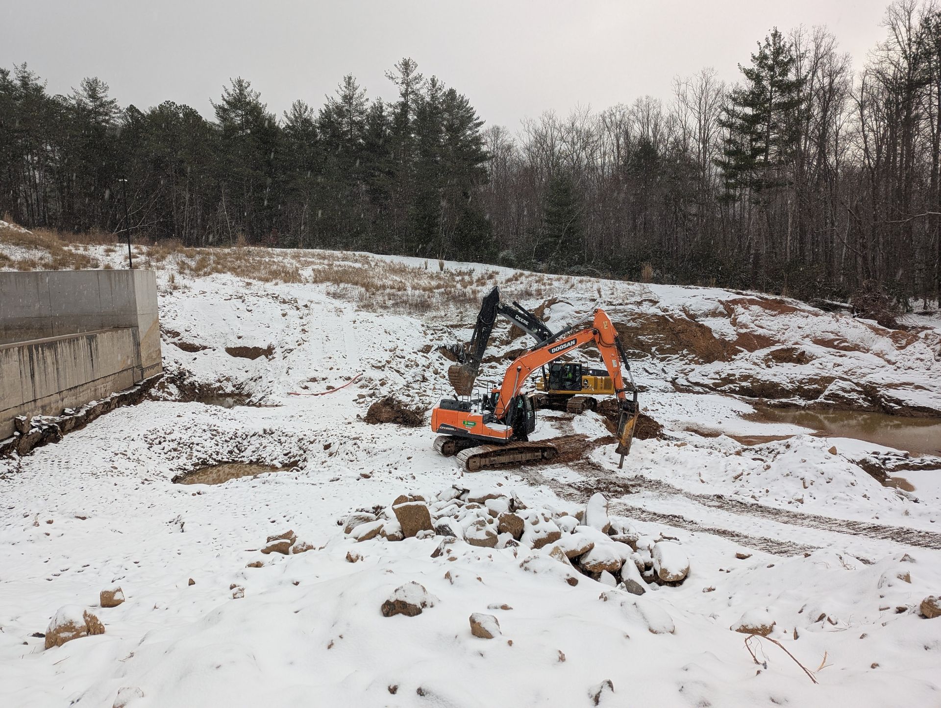 Two excavators on snowy ground, near a concrete structure, with trees in the background.