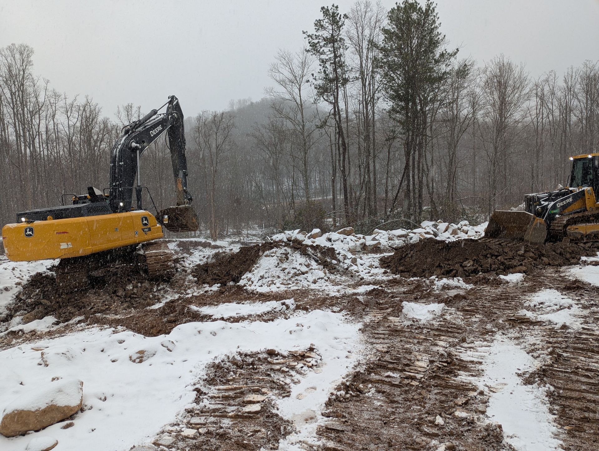 Excavator and bulldozer clearing land in snowy conditions, near a forested area.
