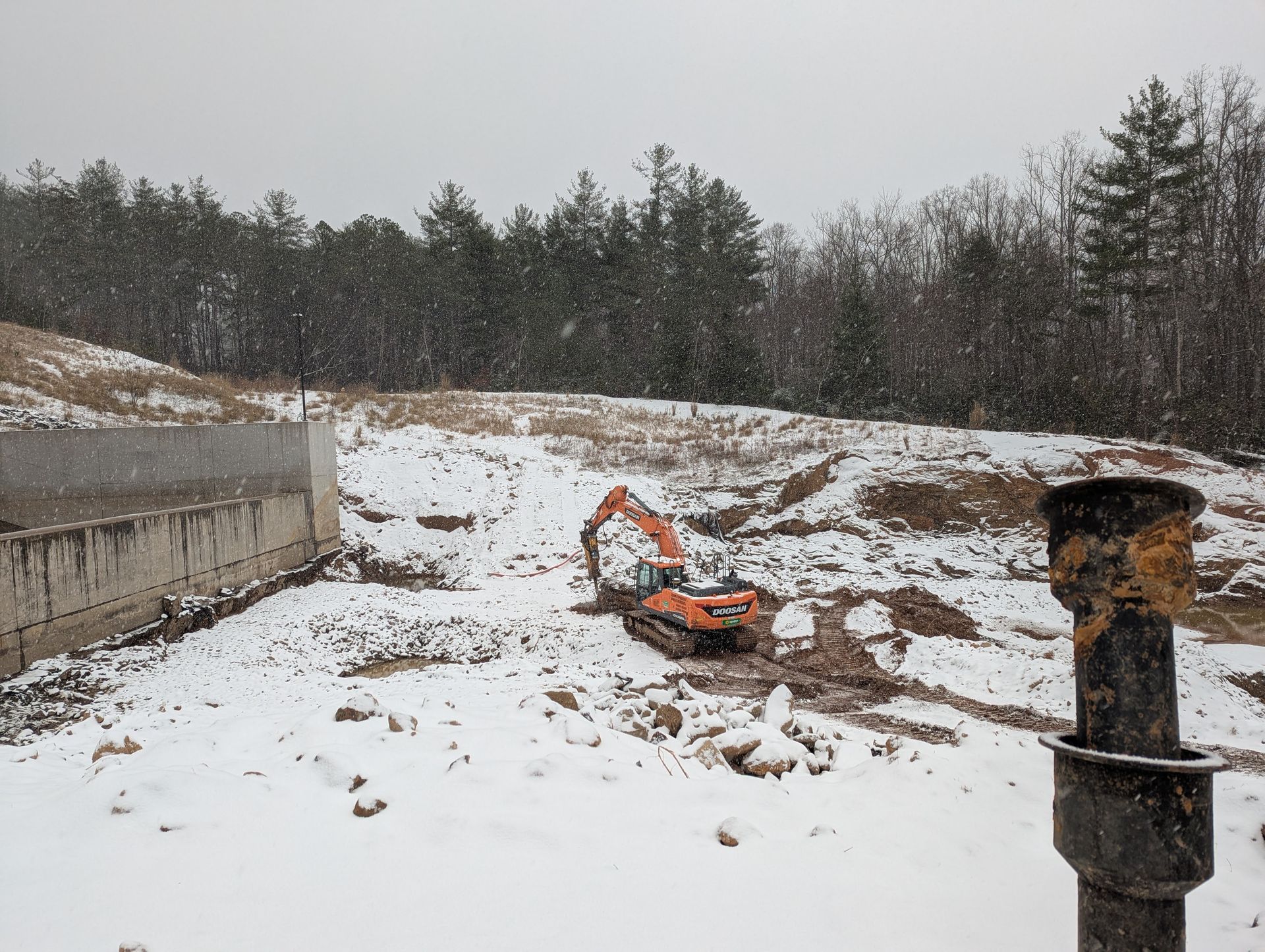 An excavator works in a snowy construction site near a concrete wall and trees.