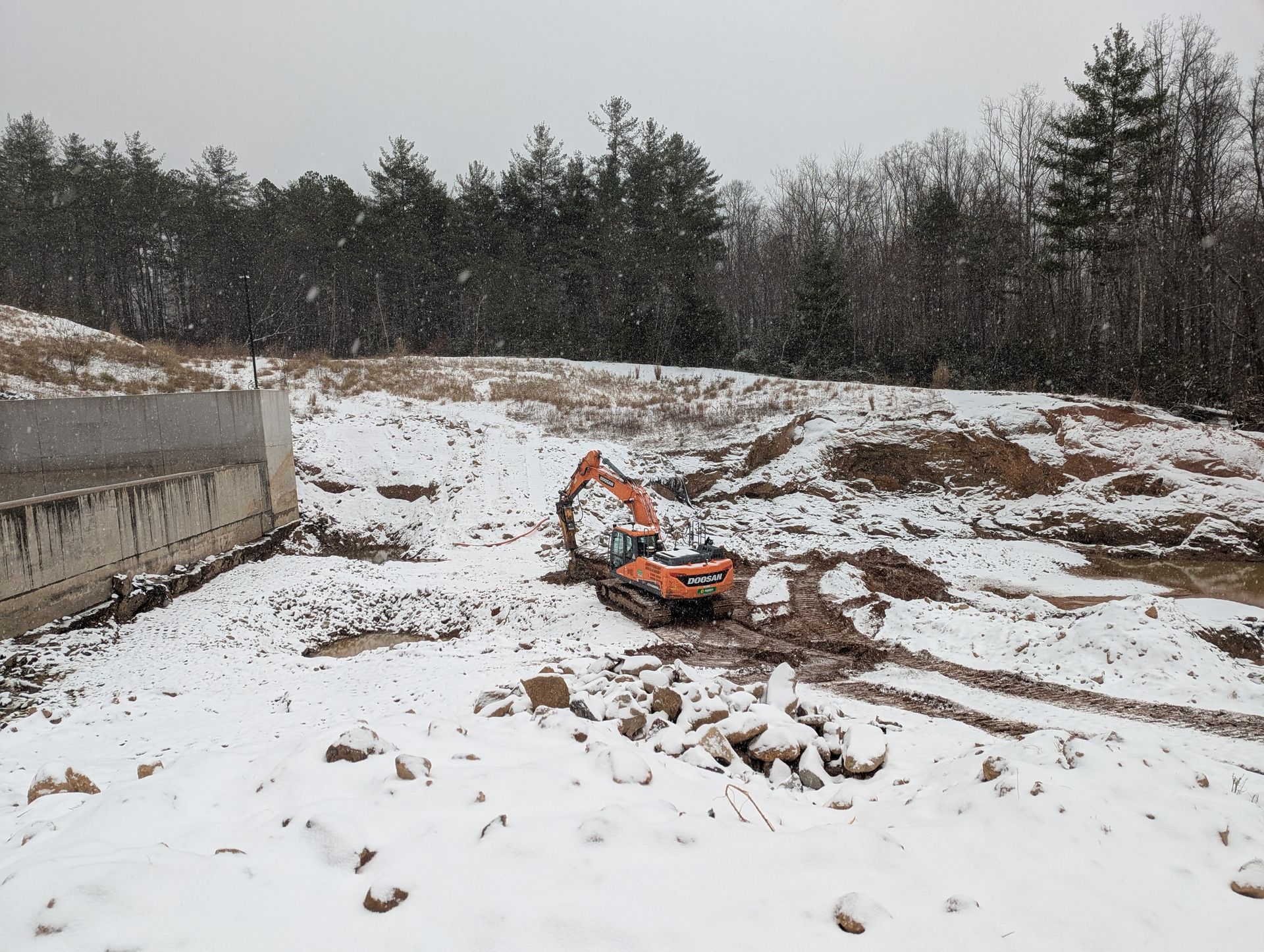 Excavator digging in snowy construction site near concrete wall and treeline.