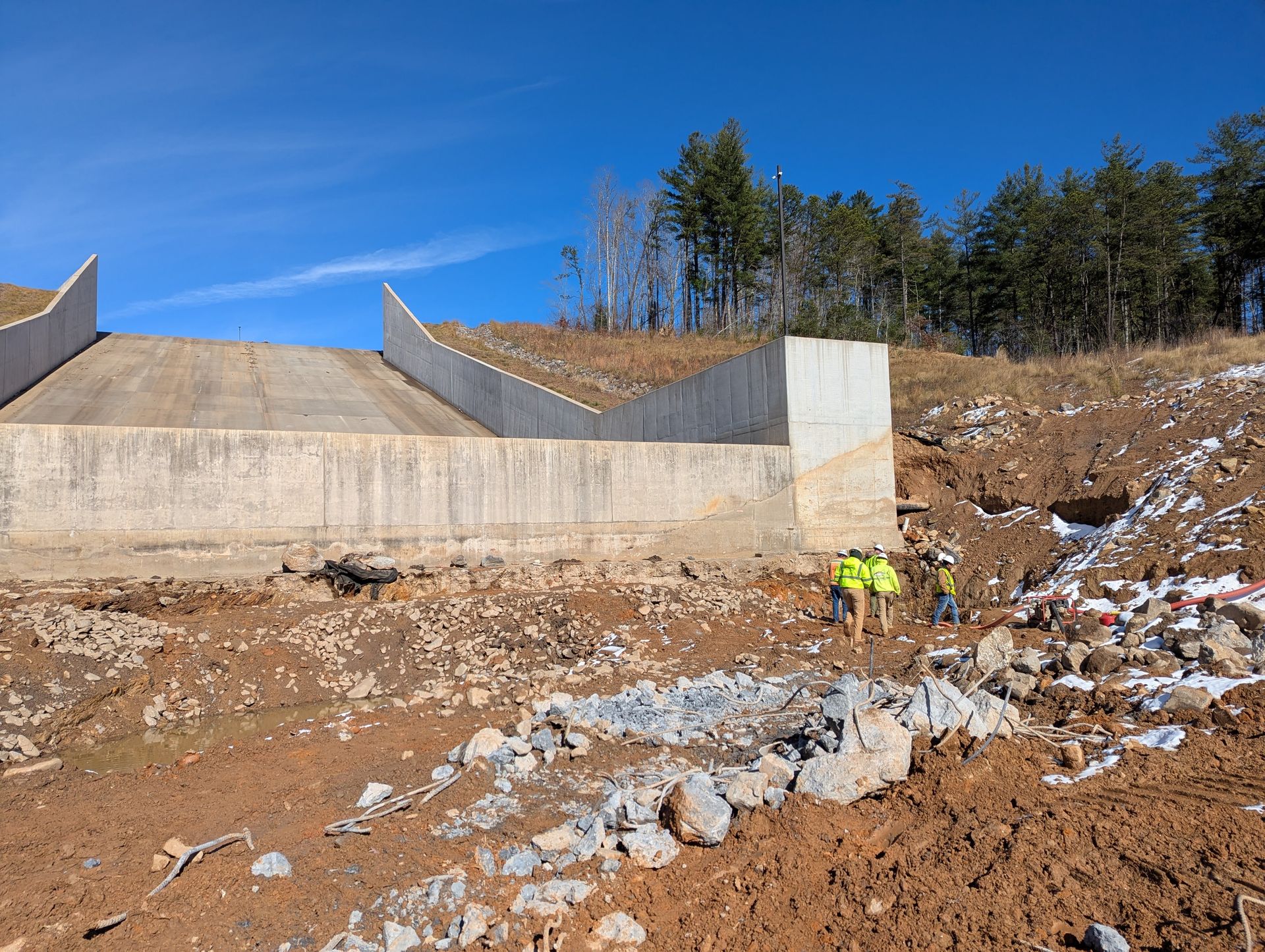 Construction site. Workers in safety vests by a concrete retaining wall on a hillside under a blue sky.