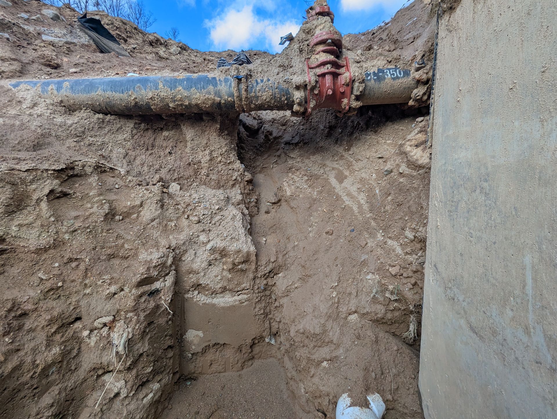 Trench exposes water pipe with red valve; soil and concrete walls.